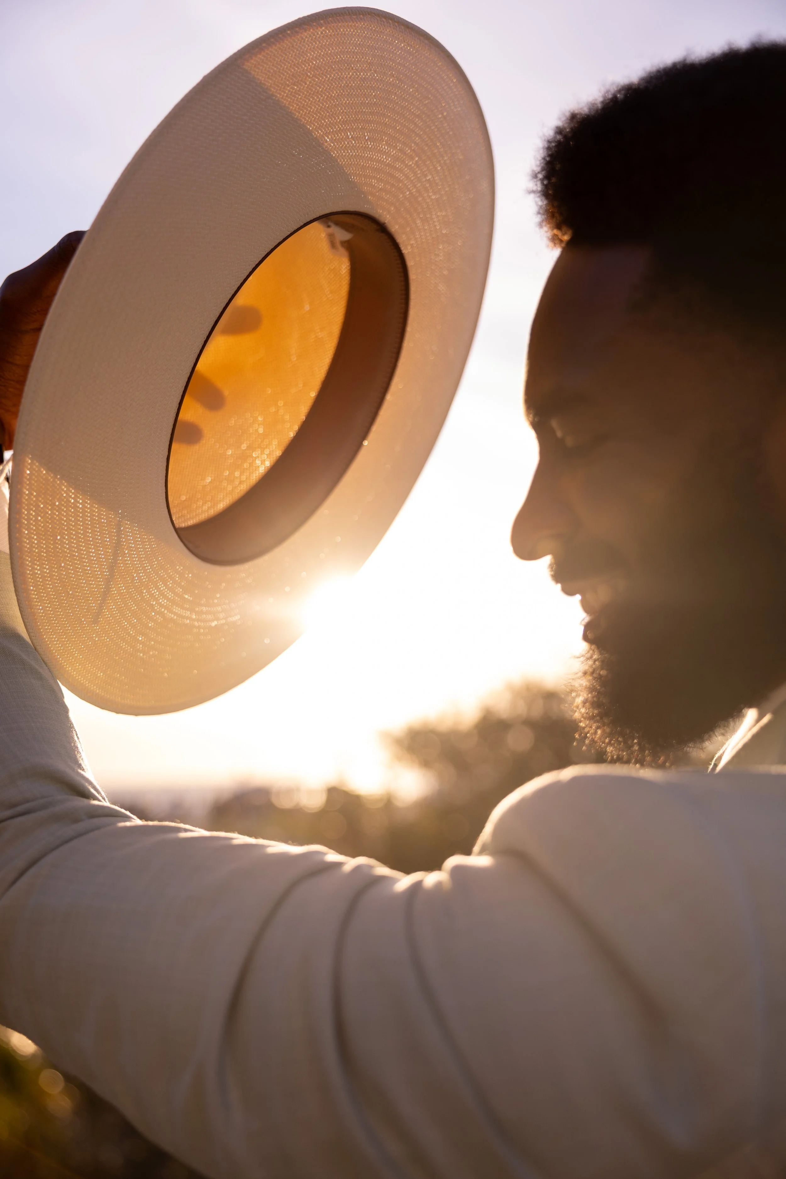 A person with dark skin and short curly hair holding a wide-brimmed hat with a yellow inner lining up to their face, with sunlight behind them creating a warm glow.