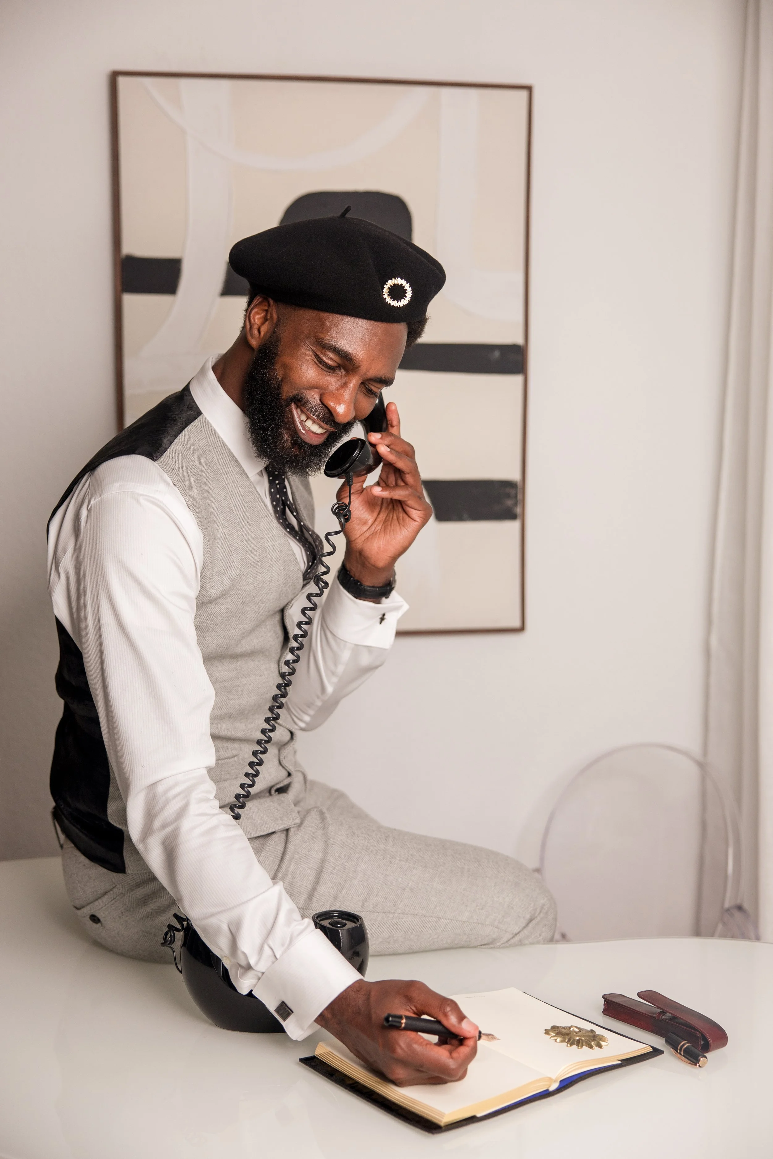 A man with a beard, wearing a black beret and a gray vest, sitting cross-legged on a white table, smiling and talking on a landline phone, with a black globe, a pen, a notebook, and writing materials on the table, in front of an abstract framed art piece on the wall.
