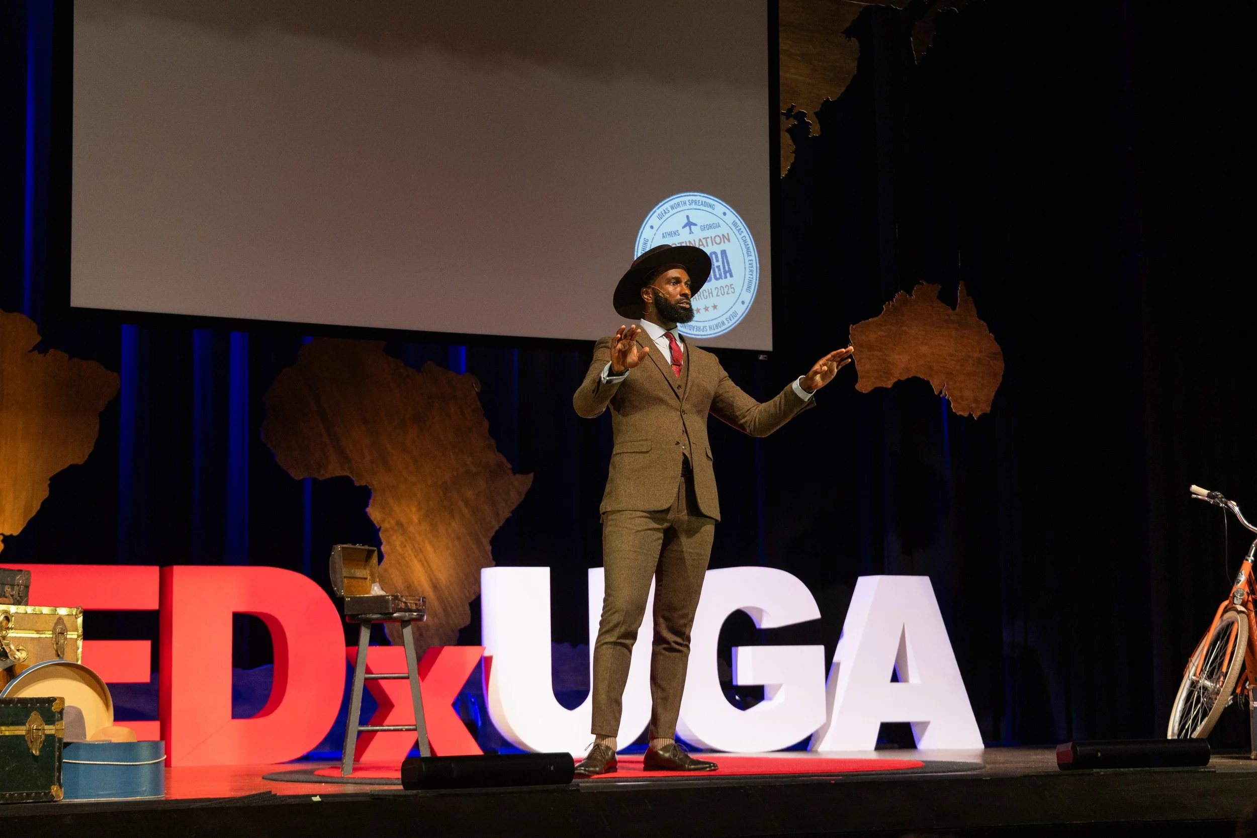 A man dressed in a suit and hat giving a presentation on a stage with large TED and Google letters and a world map backdrop.