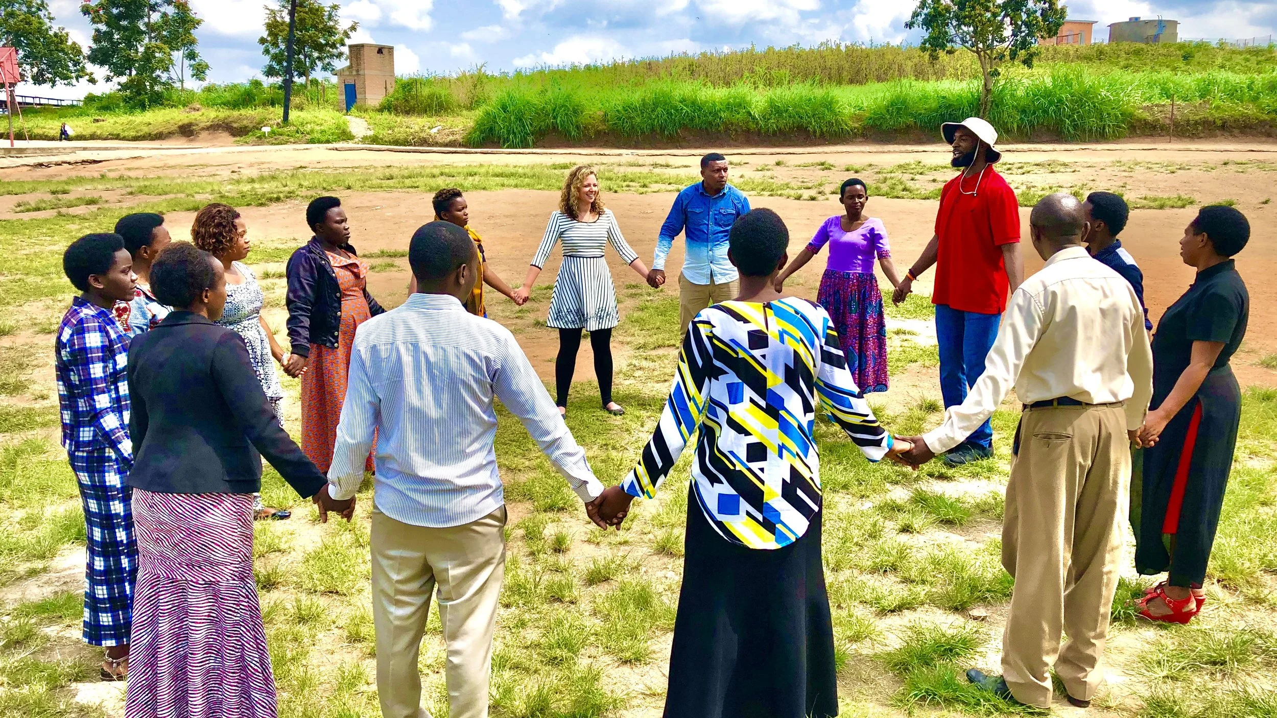 A diverse group of people holding hands in a circle outdoors on a grassy area, smiling and engaging in a communal activity.
