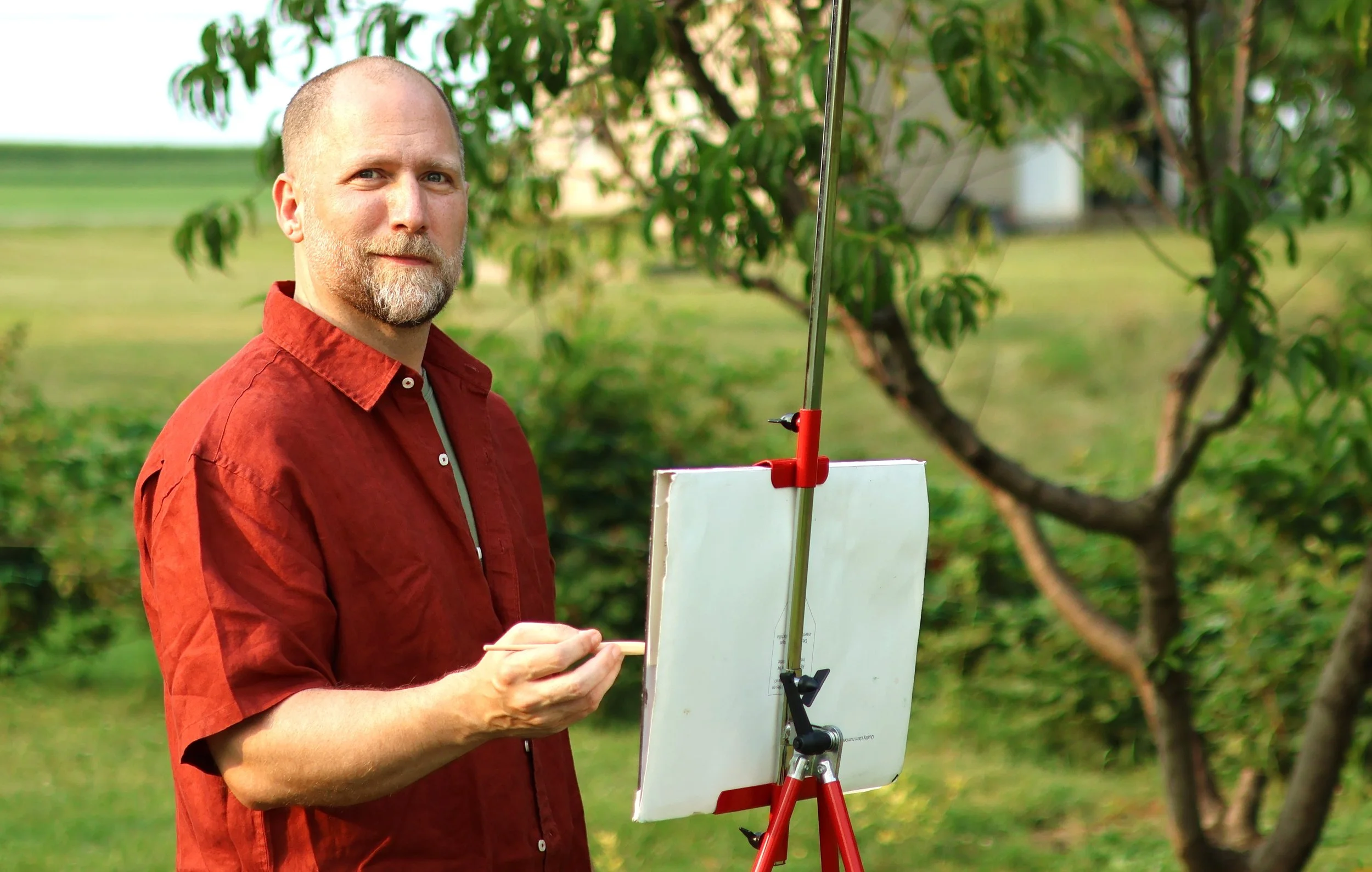 A man in a red shirt stands outdoors holding a paintbrush near an empty white canvas on an easel, with trees and greenery in the background.