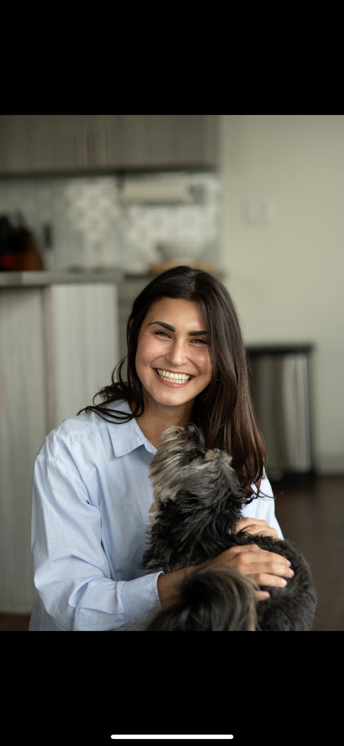 A woman with long dark hair smiling and holding a small, fluffy dog in a home interior.