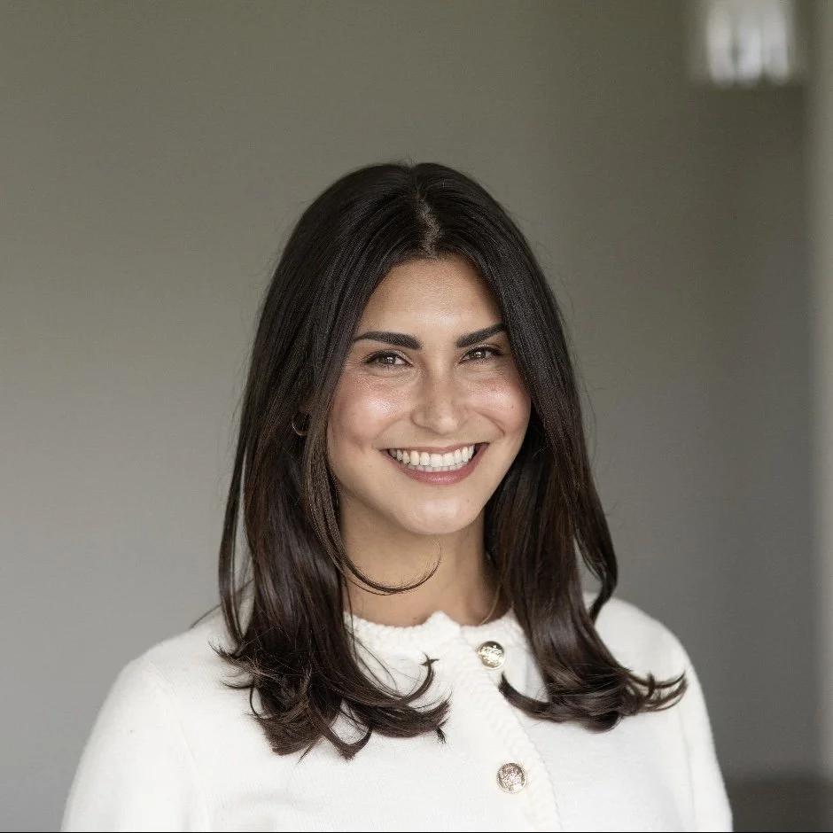 A smiling young woman with long dark hair, wearing a white top with buttons, standing against a plain gray background.