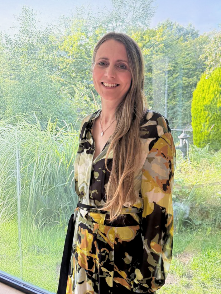 A woman with long, light brown hair smiling, standing outdoors in front of large windows with a view of a lush green garden.