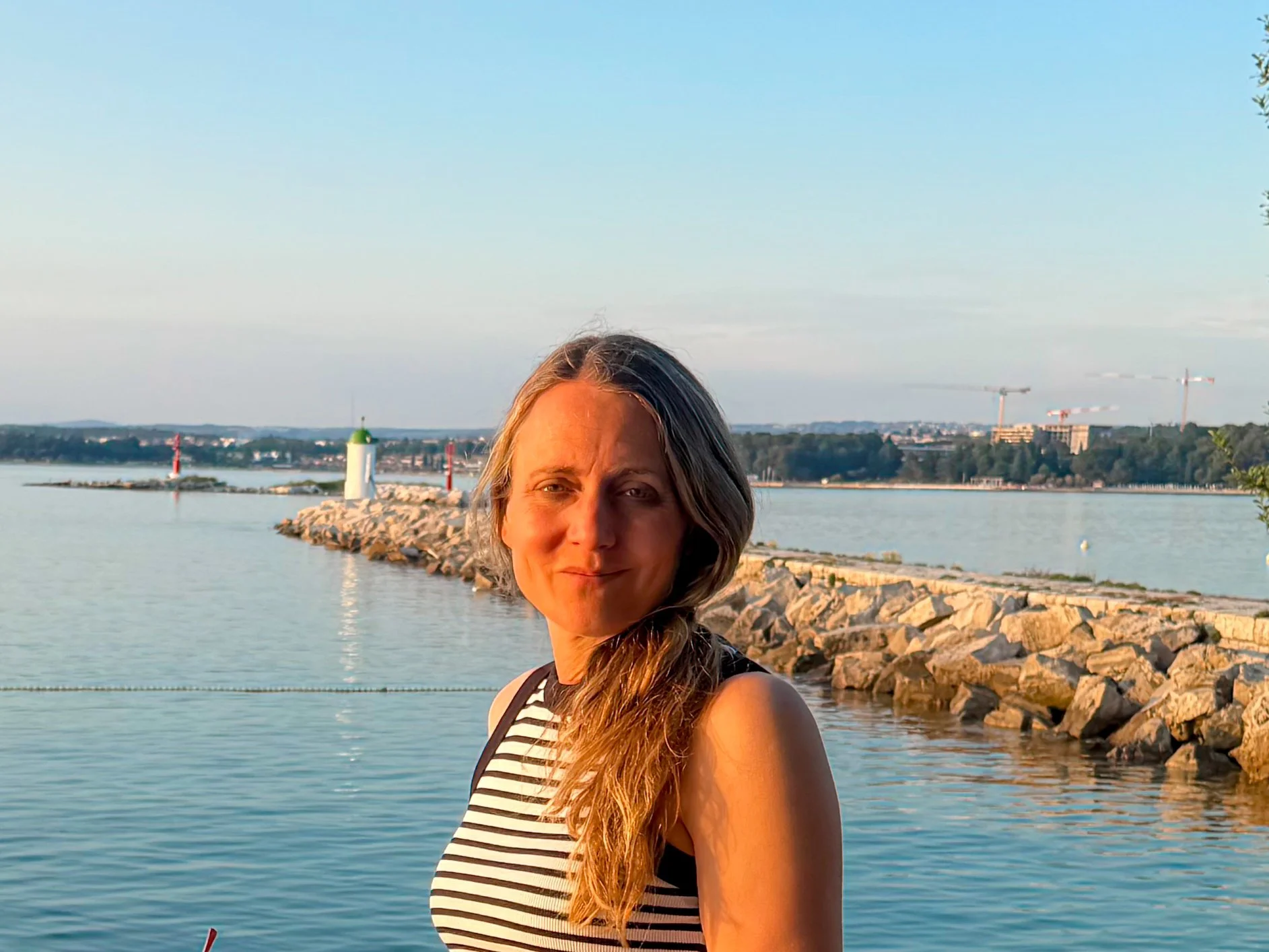 A woman with long, wavy hair wearing a black and white striped sleeveless top standing near a body of water during sunset.