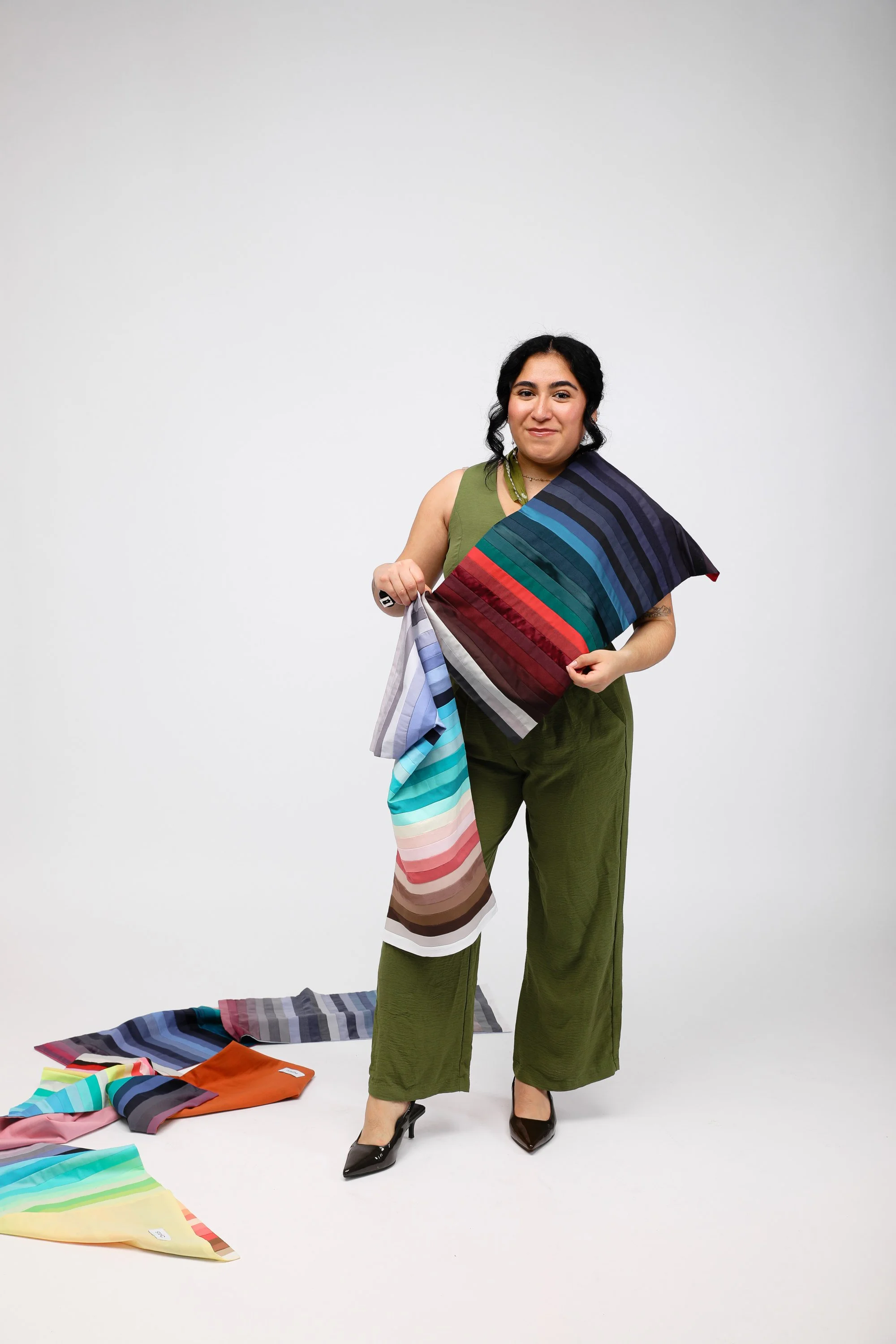A woman with dark hair and olive green outfit holding a colorful striped silk scarf and standing in front of a plain white background with more scarves on the floor.