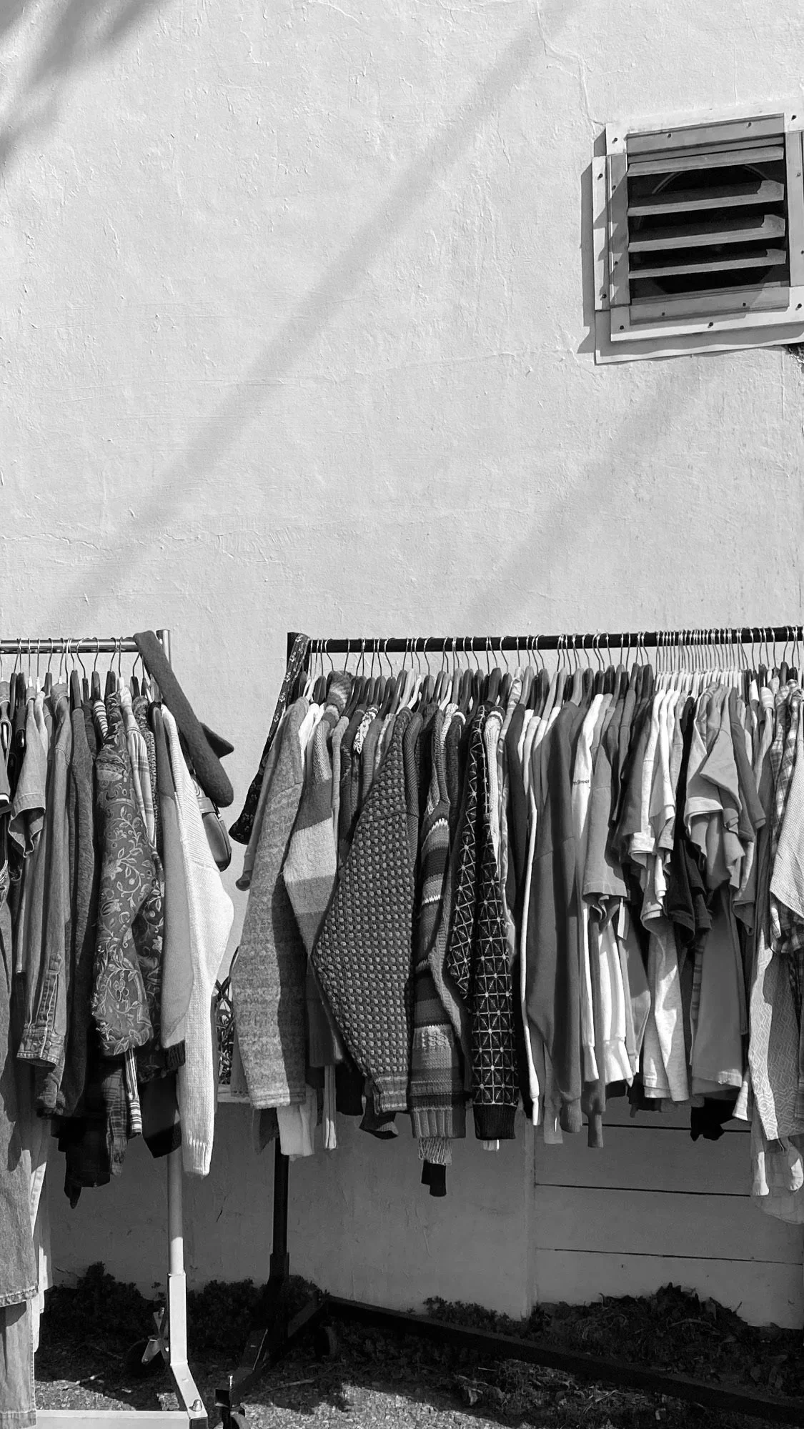 Clothes hanging on two racks outside near a white wall with a small ventilation window