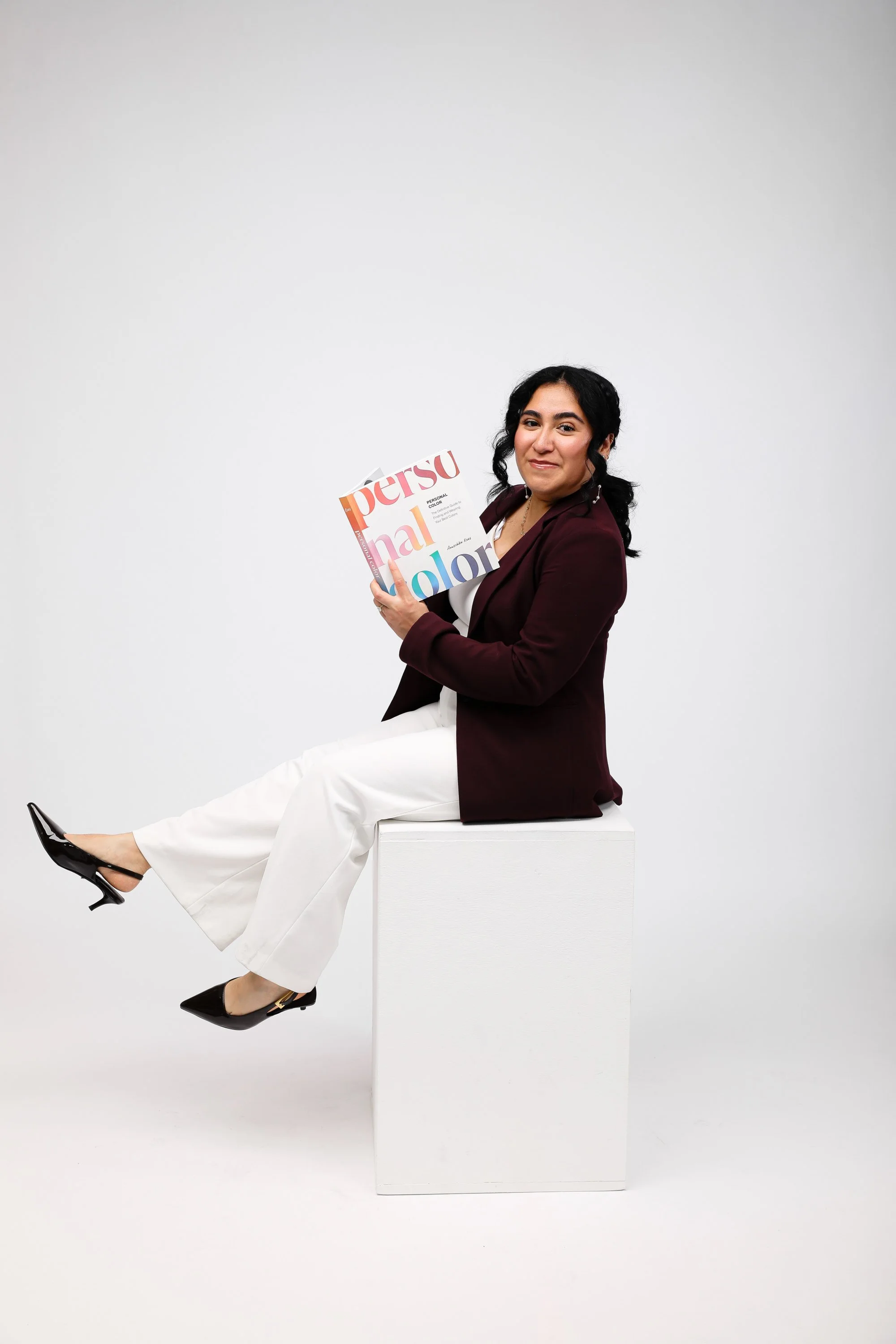 A woman sitting on a white block, holding a fashion magazine titled 'Personal Color,' with a plain white background.