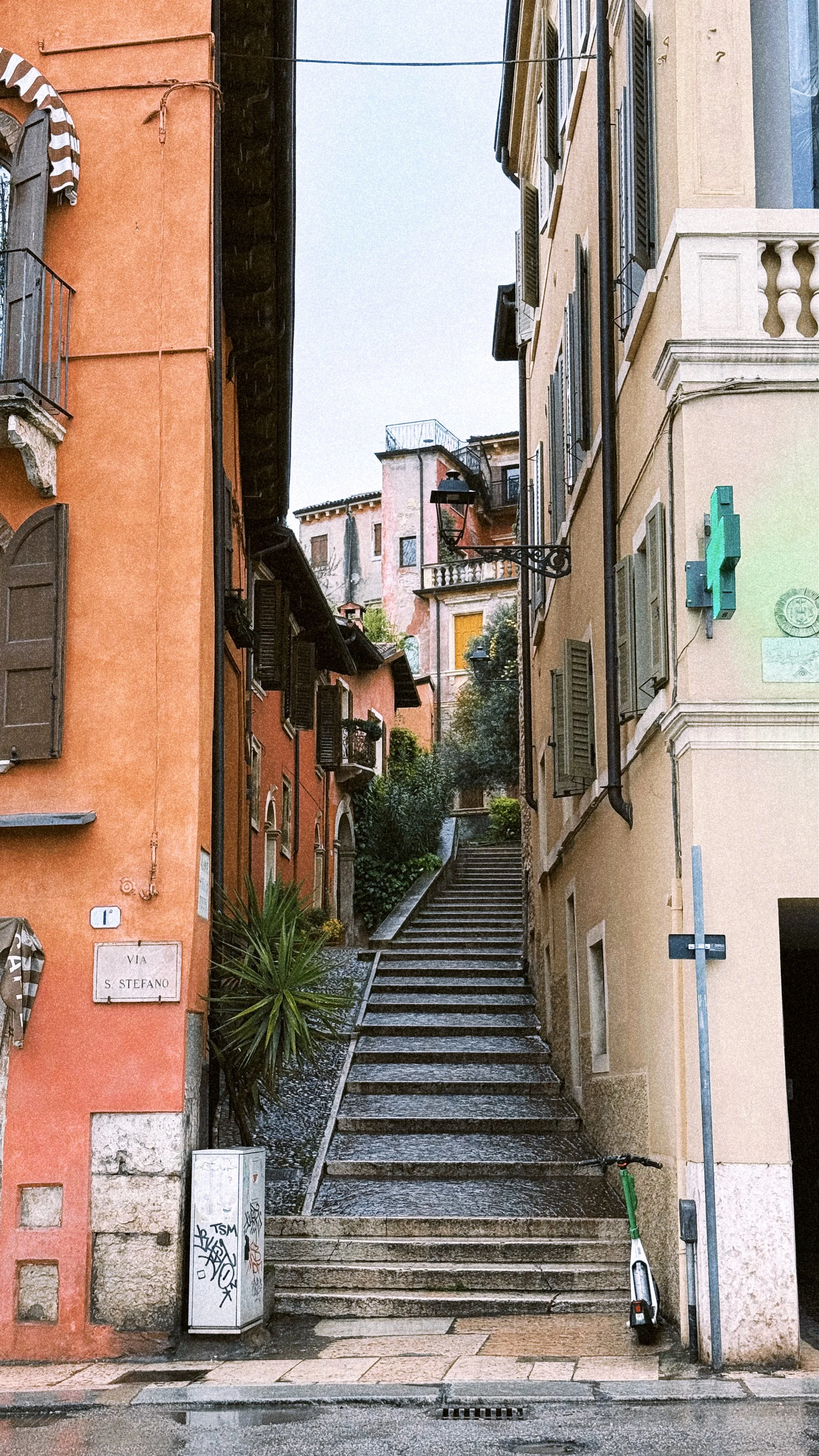 A narrow street with stairs leading upward between colorful buildings in an Italian town. The left building is orange with brown shutters and a small balcony. The right building is pale yellow with shutters and a sidewalk with a scooter parked against the wall. Vegetation is visible further up the stairs.