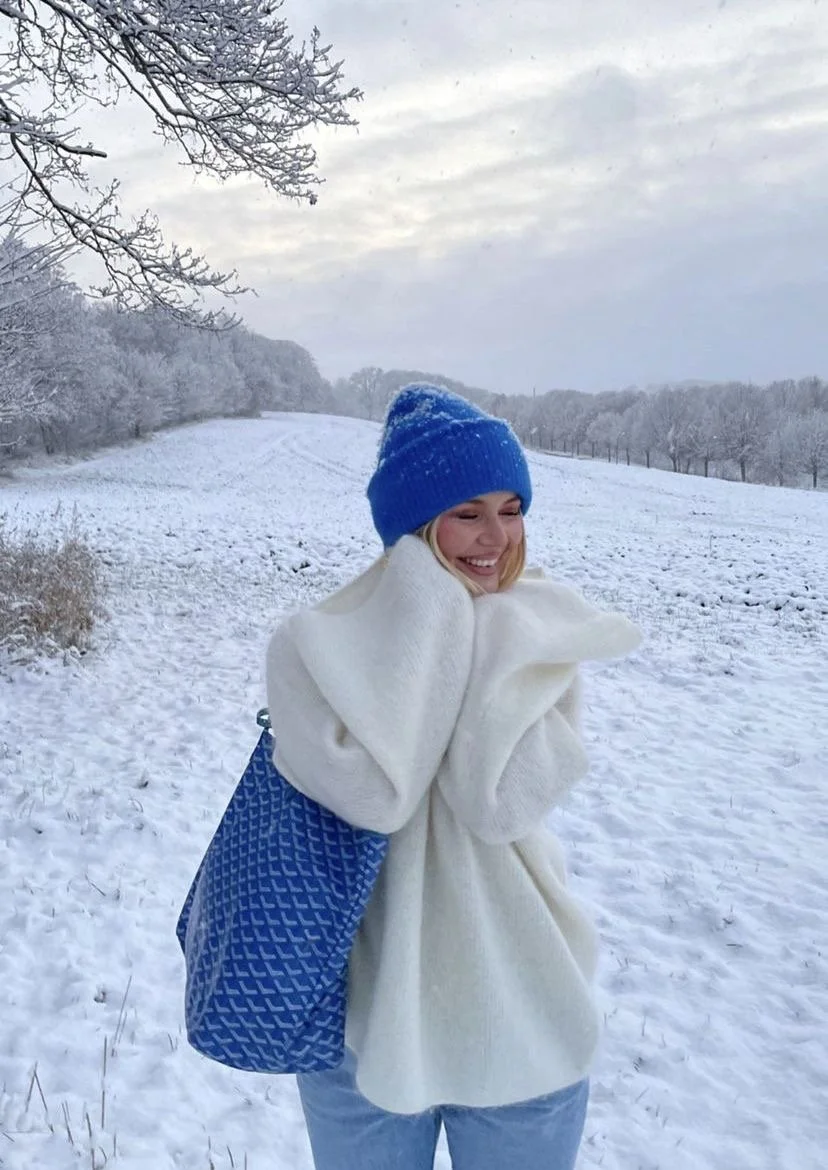 A woman dressed warmly in a white coat and blue knit hat, standing in a snowy outdoor landscape, smiling and holding her face with both hands, with snow-covered trees and a cloudy sky in the background.