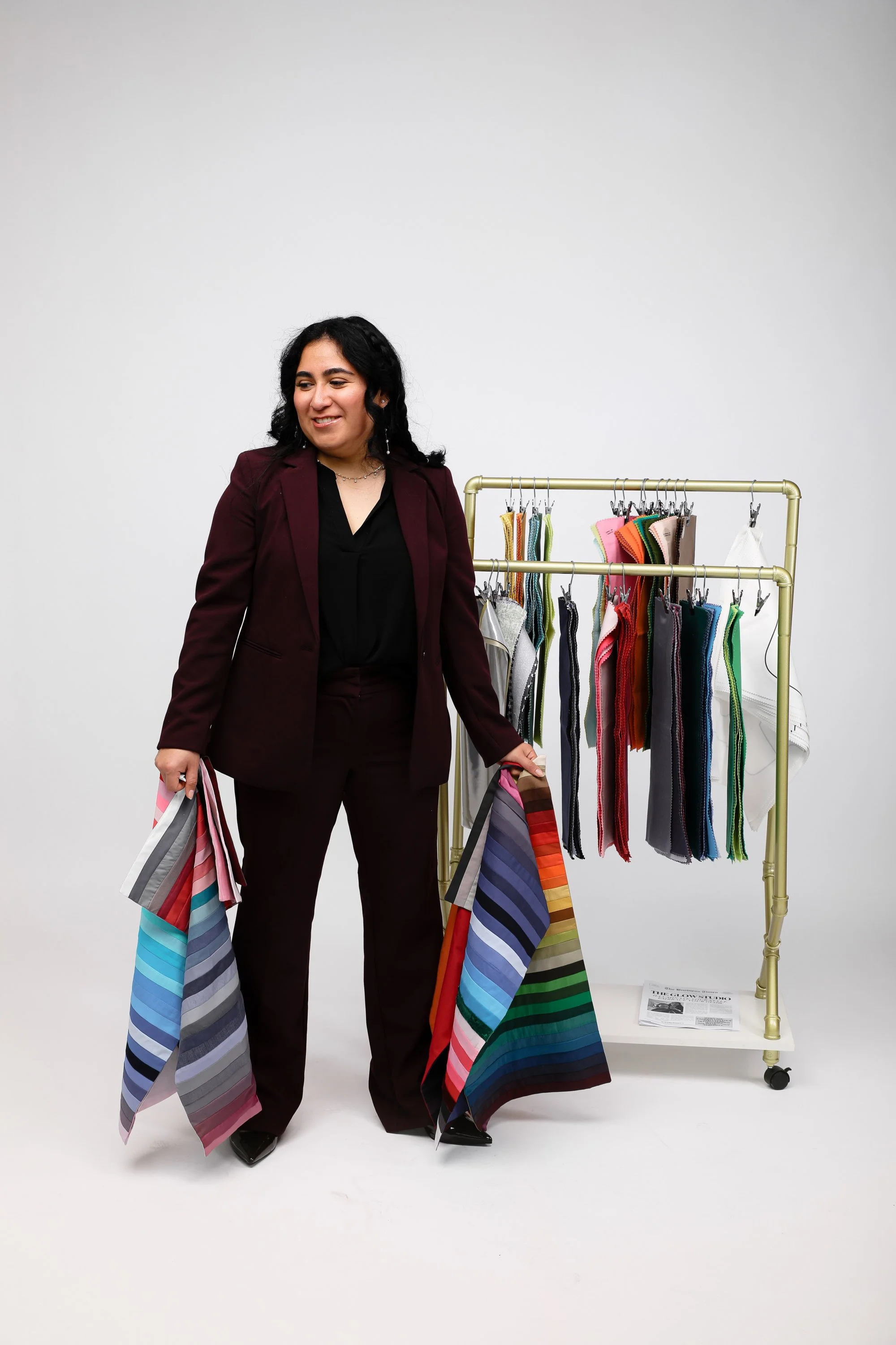 Woman in a dark red suit holding colorful fabric samples standing next to a clothing rack with various fabric swatches.