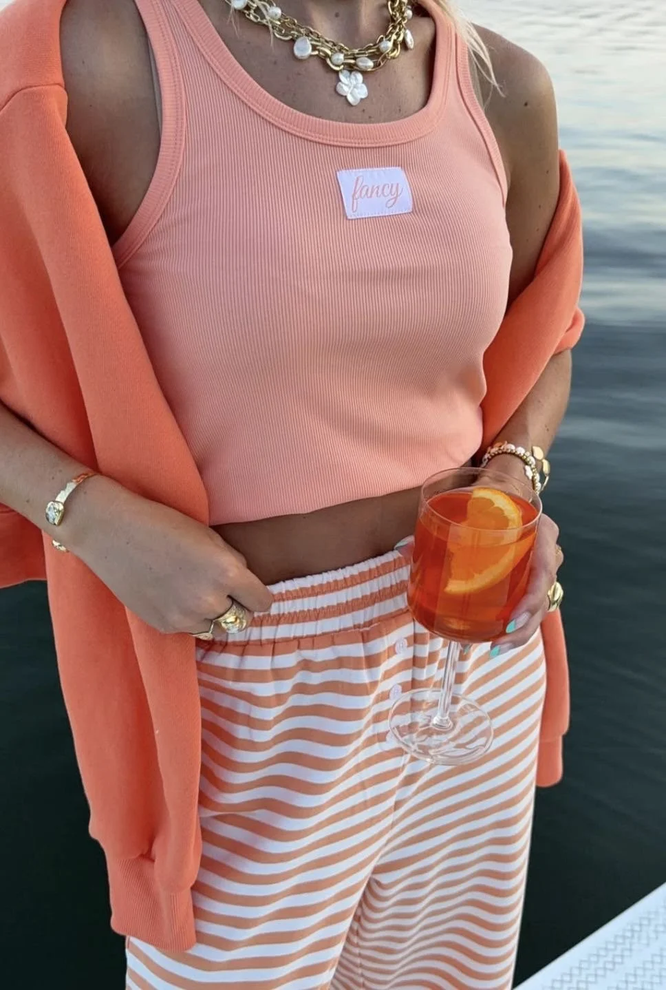 Close-up of a woman wearing a peach-colored tank top with a 'fancy' sticker, striped orange and white pants, and layered jewelry, holding a glass of orange-colored drink with orange slices, standing near water.