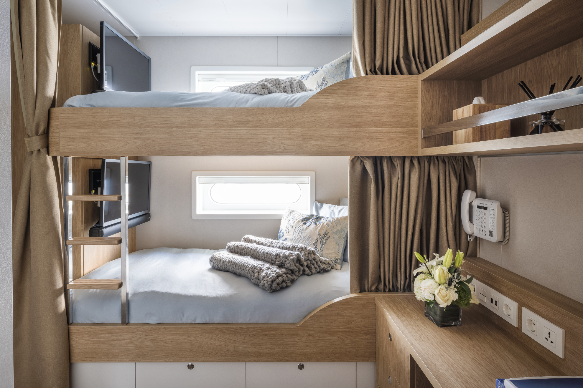 Two beds in a compact room with wooden frames, pillows, blankets, and a small window between them, with a TV mounted on the wall near the top bed and a bedside table with a flower arrangement, power outlets, and a telephone.