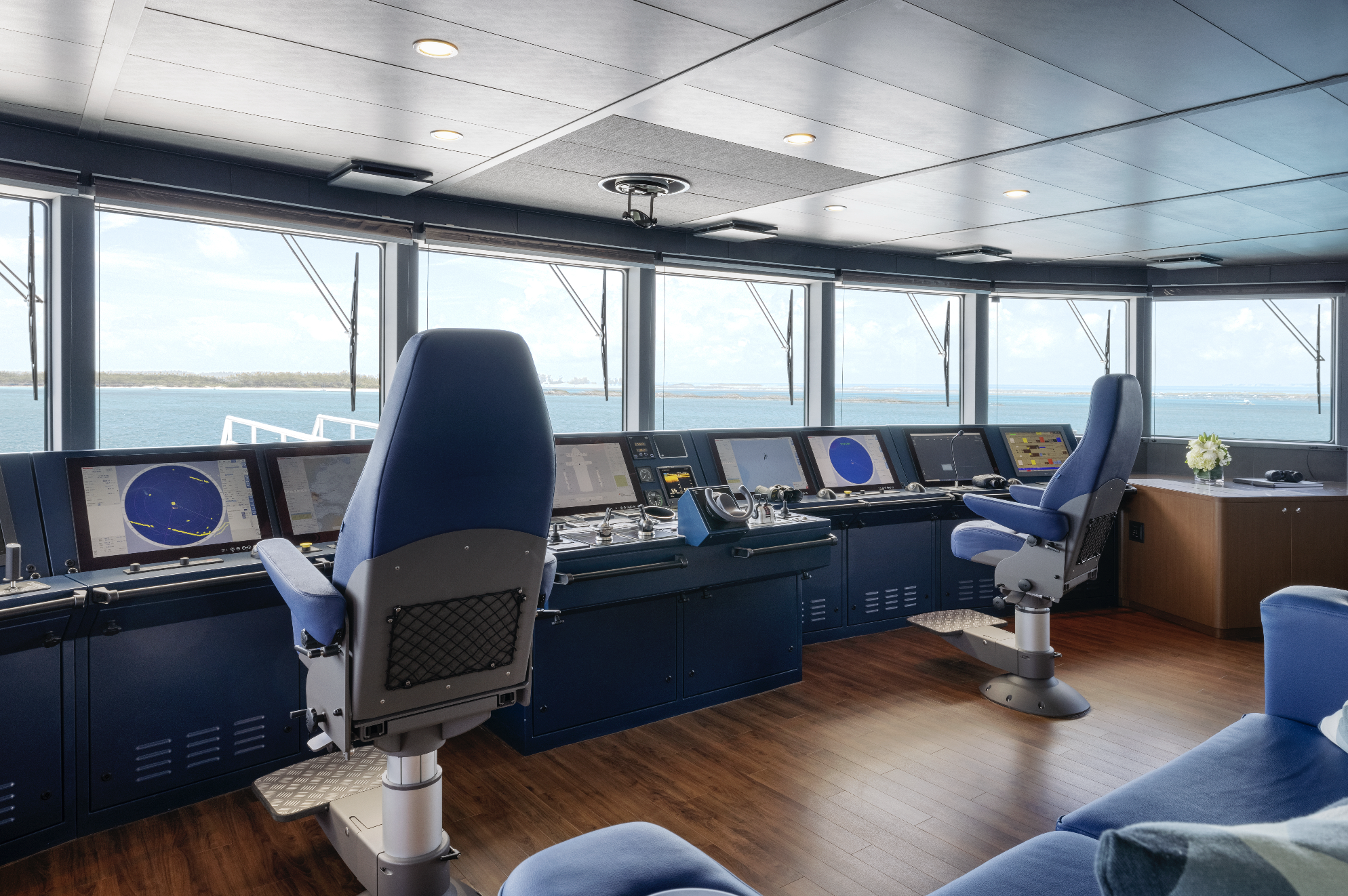 Bridge of a ship with multiple control monitors and chairs, overlooking the water outside through large windows.