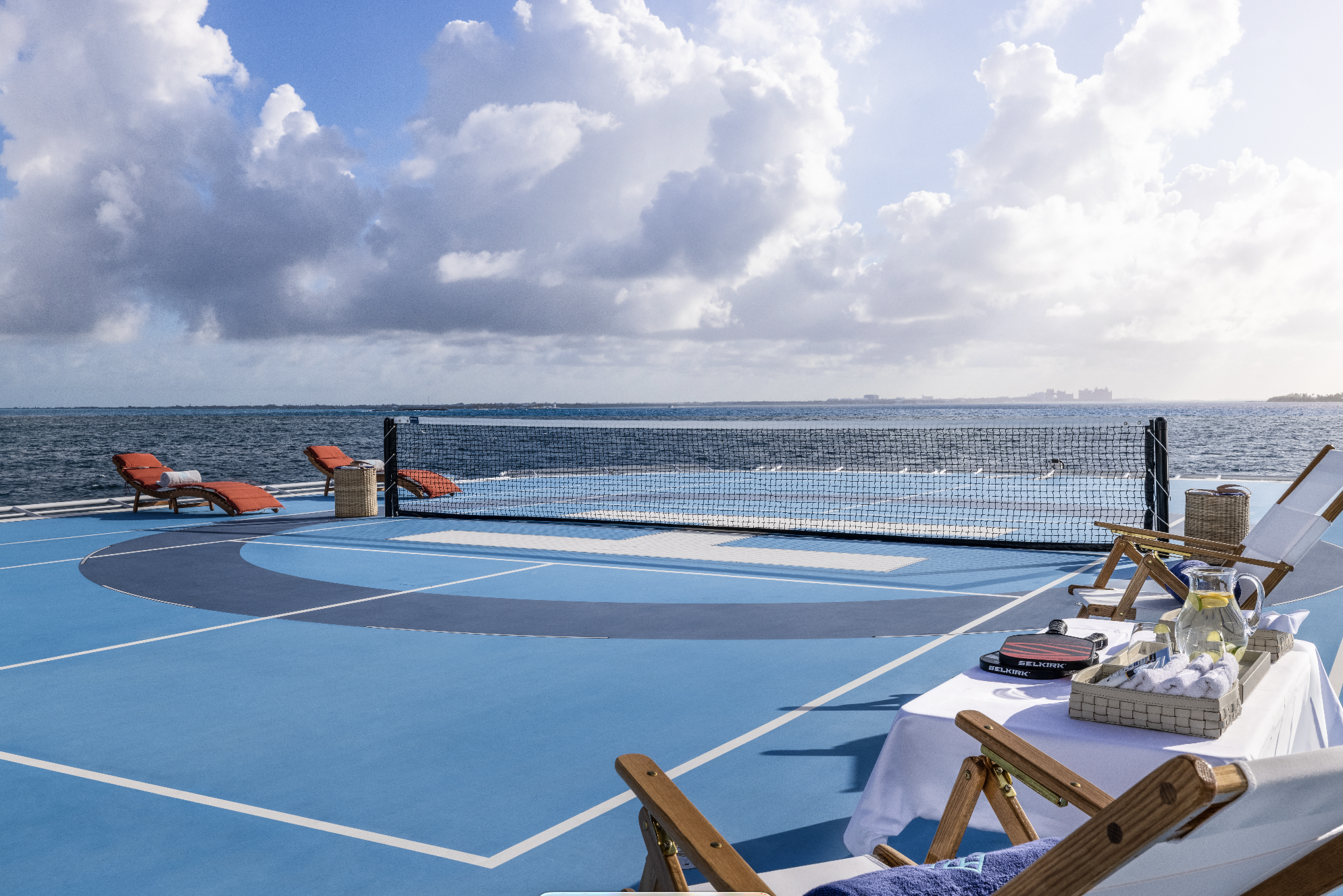 A rooftop sports court near the ocean, with lounge chairs, a table with drinks, and a volleyball net, under a partly cloudy sky.