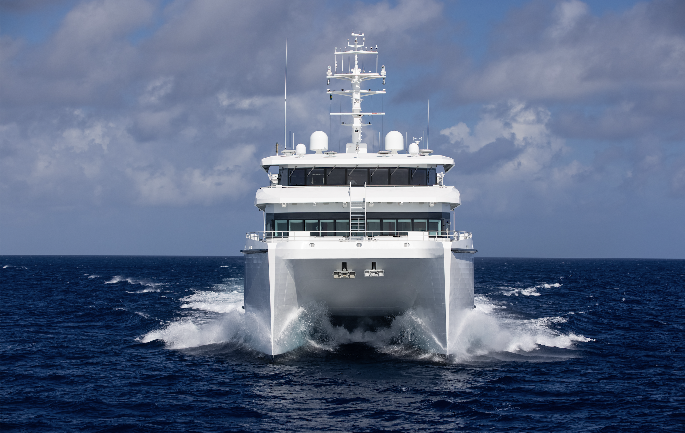 A large white yacht sailing on the open ocean, heading towards the camera with waves crashing at the bow, under a partly cloudy sky.