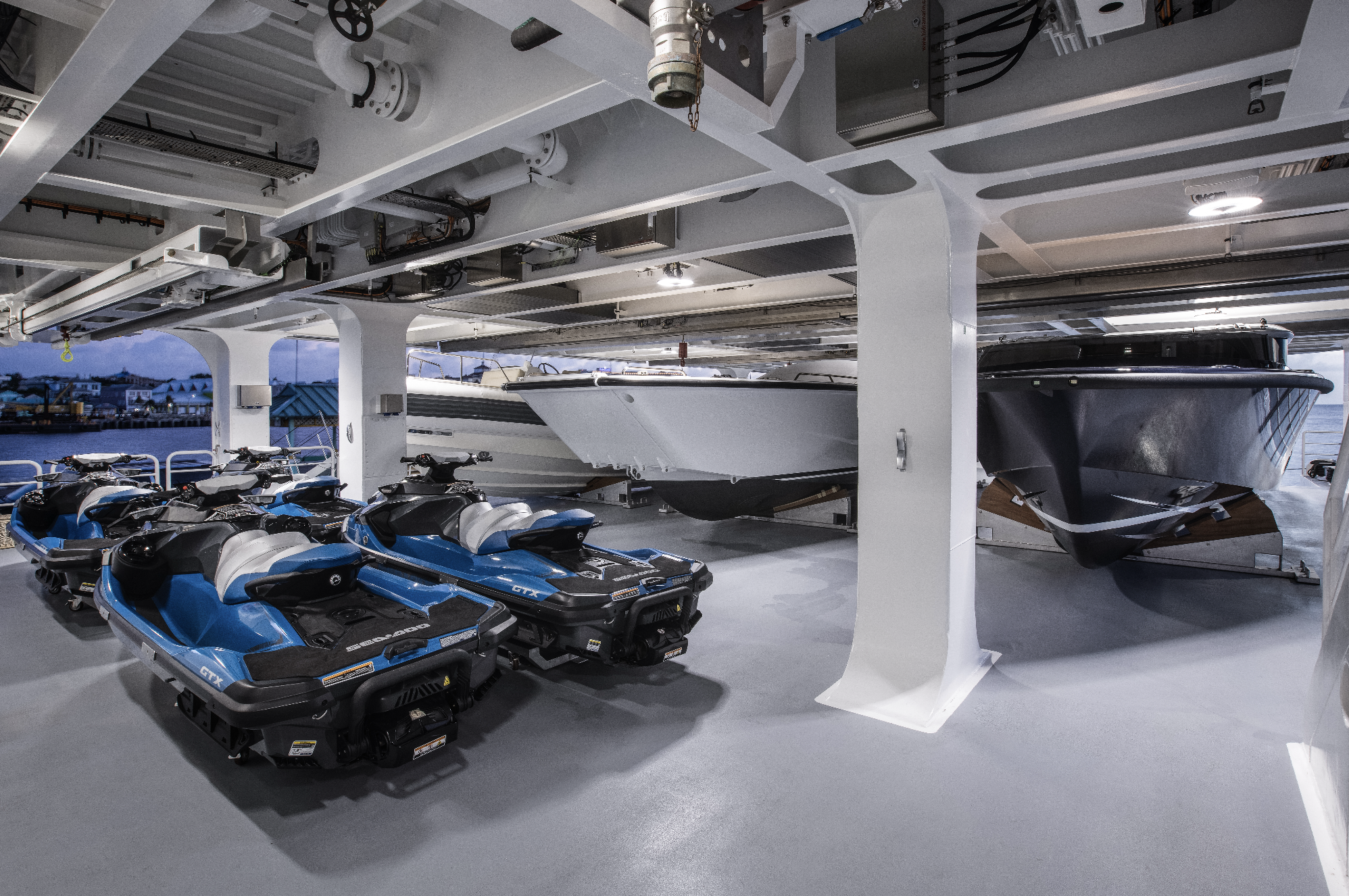 Inside a boat carrier with four jet skis, two large boats on a hydraulic lift, and a view of water and buildings outside.