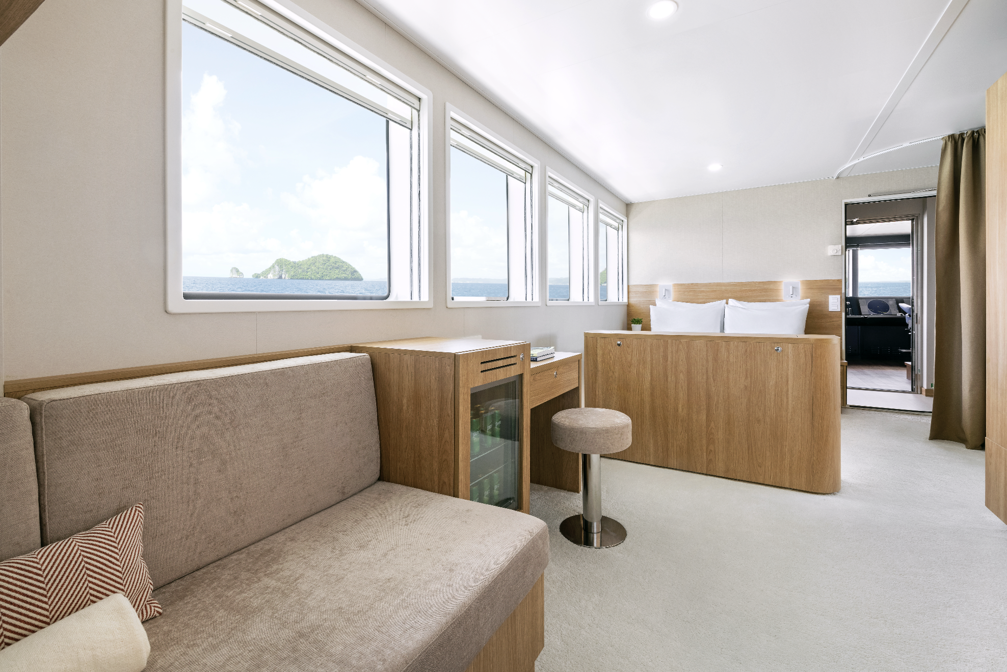 Modern hotel room with large windows, ocean view, beige sofa, wooden furniture, bed with white linens, small stool, and a door to a balcony or another room.