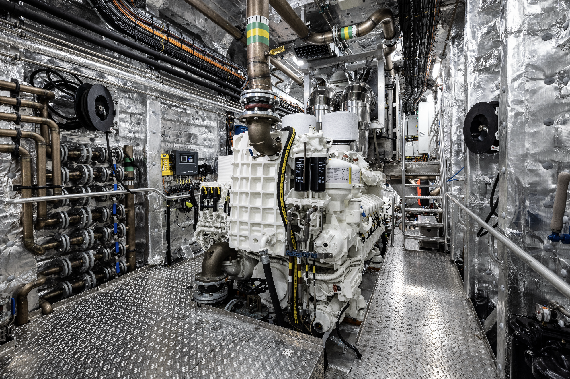 Industrial machinery and piping inside a metallic room, including a large white engine, control panels, valves, and metallic platforms.