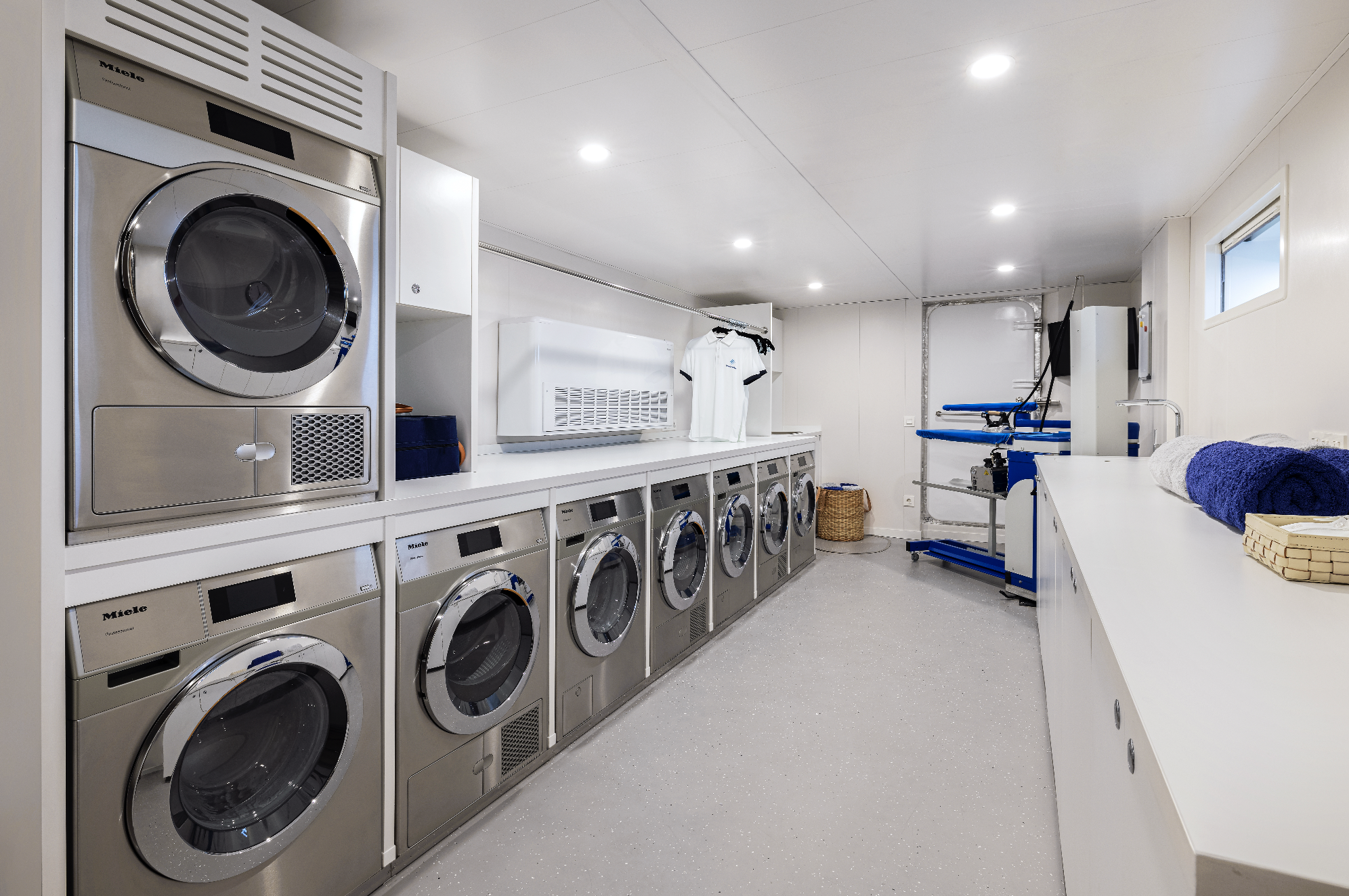 A clean laundry room with multiple front-loading wash machines and dryers, white countertops, a laundry basket, folded towels, and a utility platform in the background.