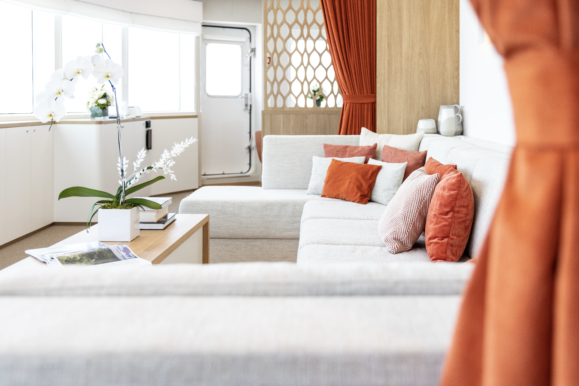 A bright living room with a white sectional sofa adorned with various orange and white pillows, a wooden coffee table with magazines and a potted orchid, large windows, and orange curtains.