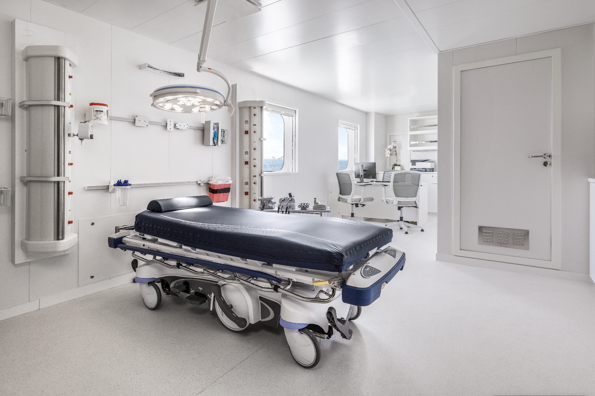 Empty hospital operating room with a medical bed, surgical light, and workstations, bright white walls, and windows showing water outside.