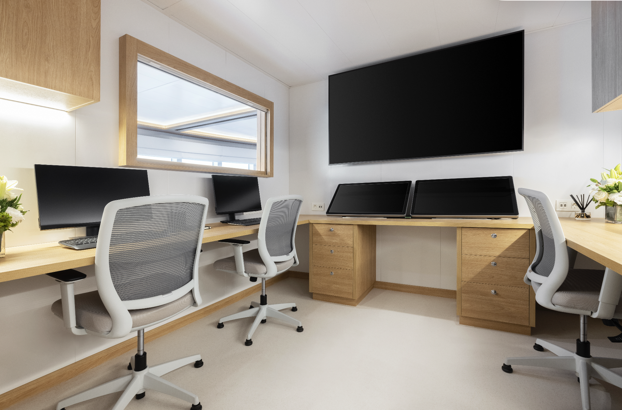 An office with three workstations, each with a computer monitor and an ergonomic chair, wooden desks, a large window, and a wall-mounted large screen.