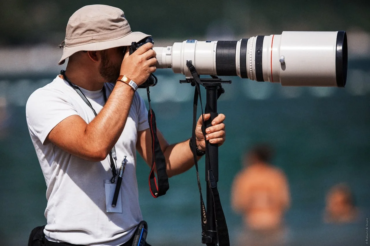 A man wearing a sun hat using a large camera lens mounted on a tripod, aiming through the lens at a distant subject, with a body of water and a few blurred figures in the background.