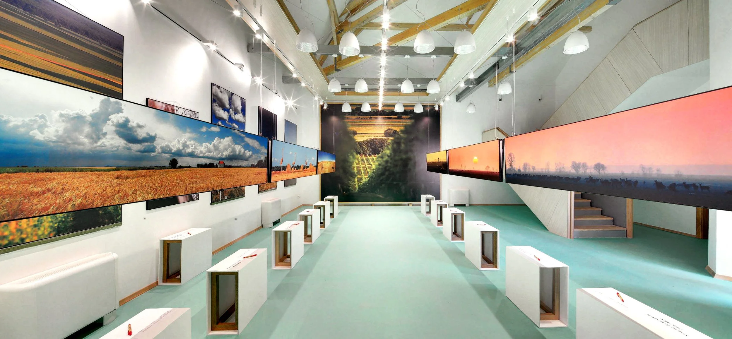 An art gallery with landscape photographs on display. The pictures include scenes of fields, clouds, sky, and sunsets, arranged in rows on white walls. The gallery has a high ceiling with visible beams and hanging lights, and white benches and displa