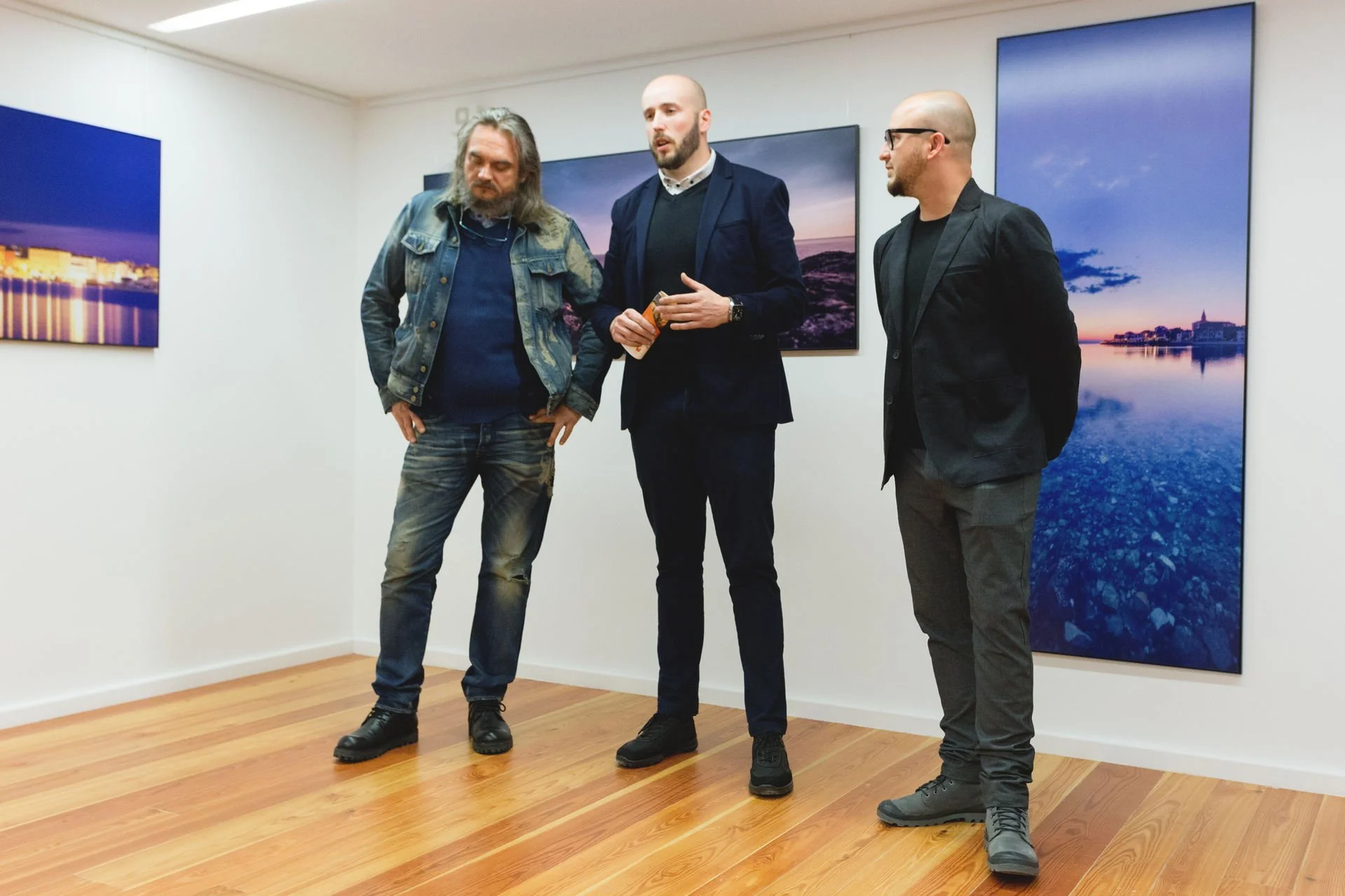 Three men standing in an art gallery, looking at photographs of landscapes on the wall.