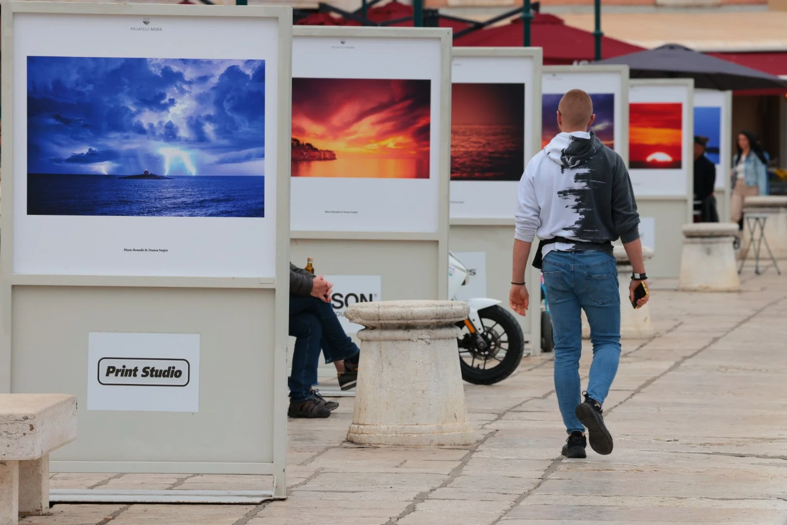 An outdoor photography exhibition displaying landscape photos of sunsets and lightning over the ocean. A man dressed in a hoodie and jeans is walking past the display, with benches and a motorcycle nearby.