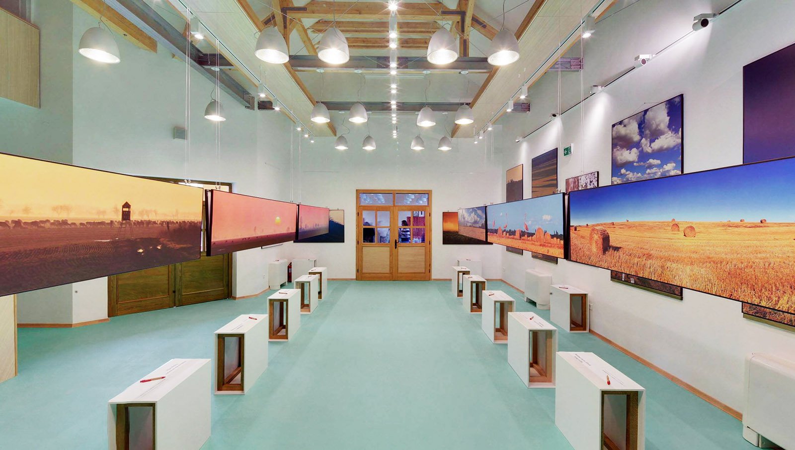 An art gallery with landscape photographs displayed on the walls. The gallery has wooden beams on the ceiling, white walls, and a light green floor. There are small white benches in front of the photographs, and a wooden door with glass panes at the 