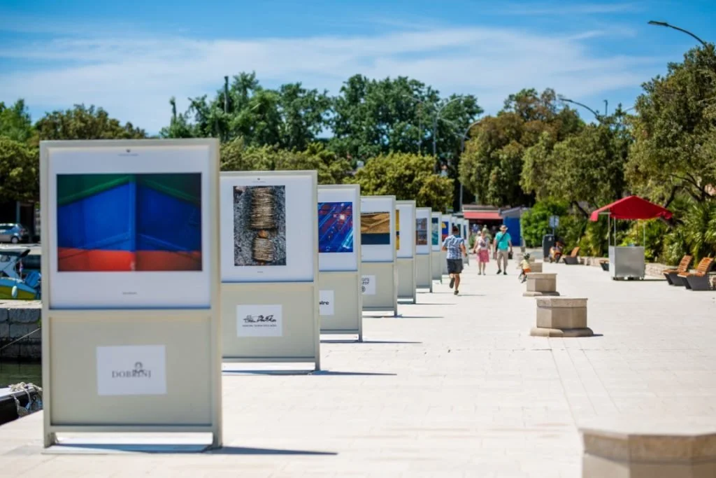 Outdoor art gallery with framed photographs displayed along a pathway, with people walking and trees in the background.
