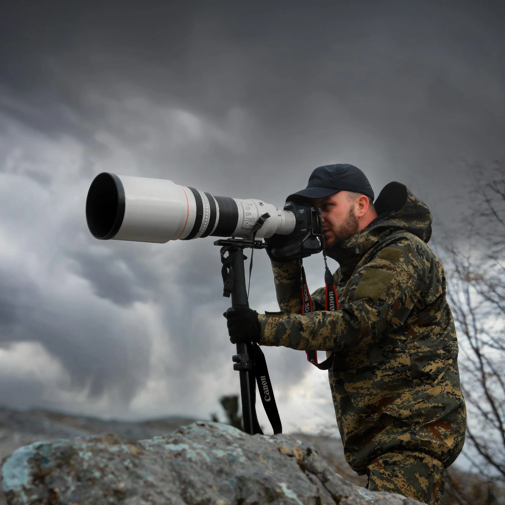 Dražen Stojčić, photographer from Osijek, Croatia. A man in camouflage jacket and black cap using a large telephoto camera lens mounted on a tripod outdoors with a cloudy sky in the background.