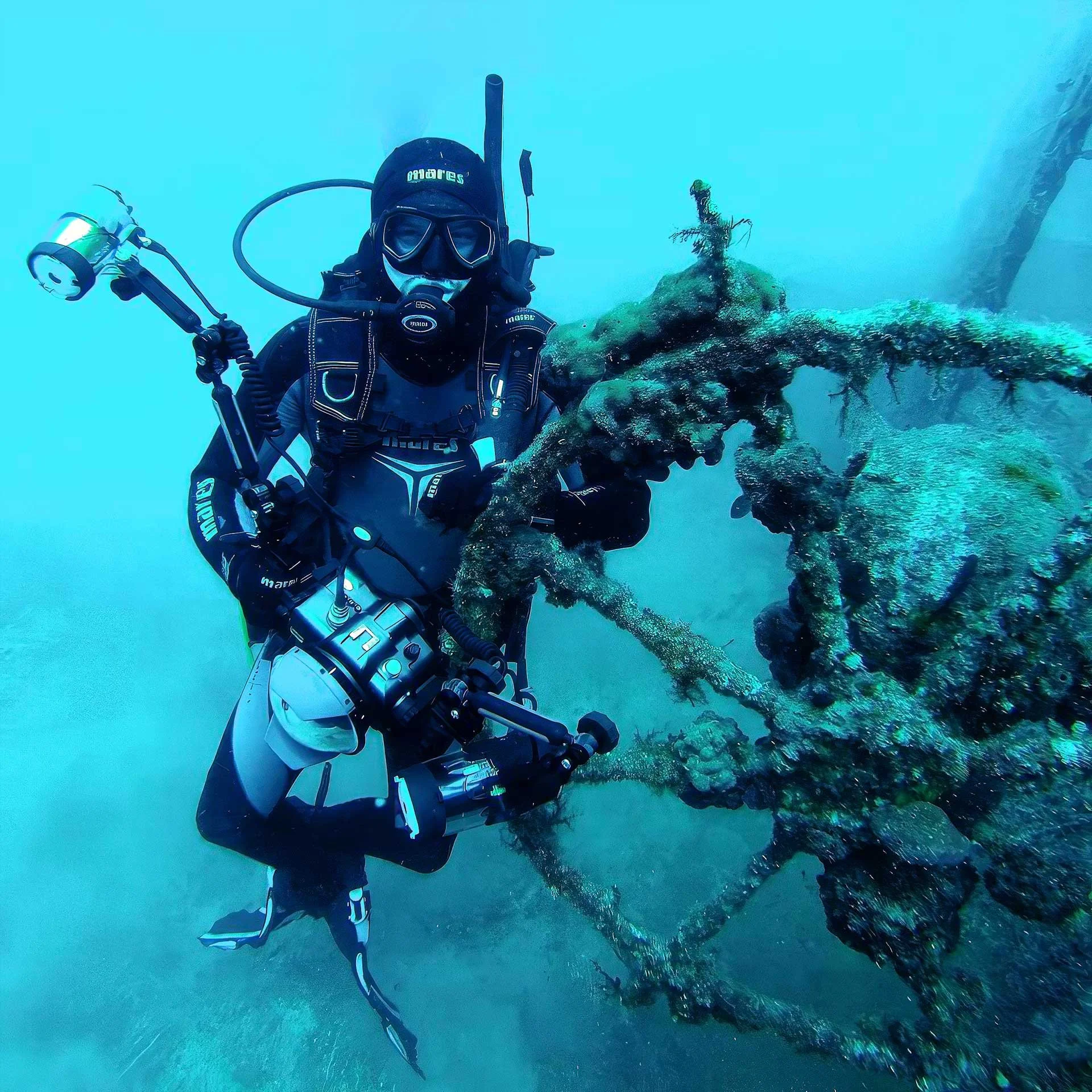 A diver in full scuba gear exploring underwater near a rusted, coral-covered structure or wreckage.