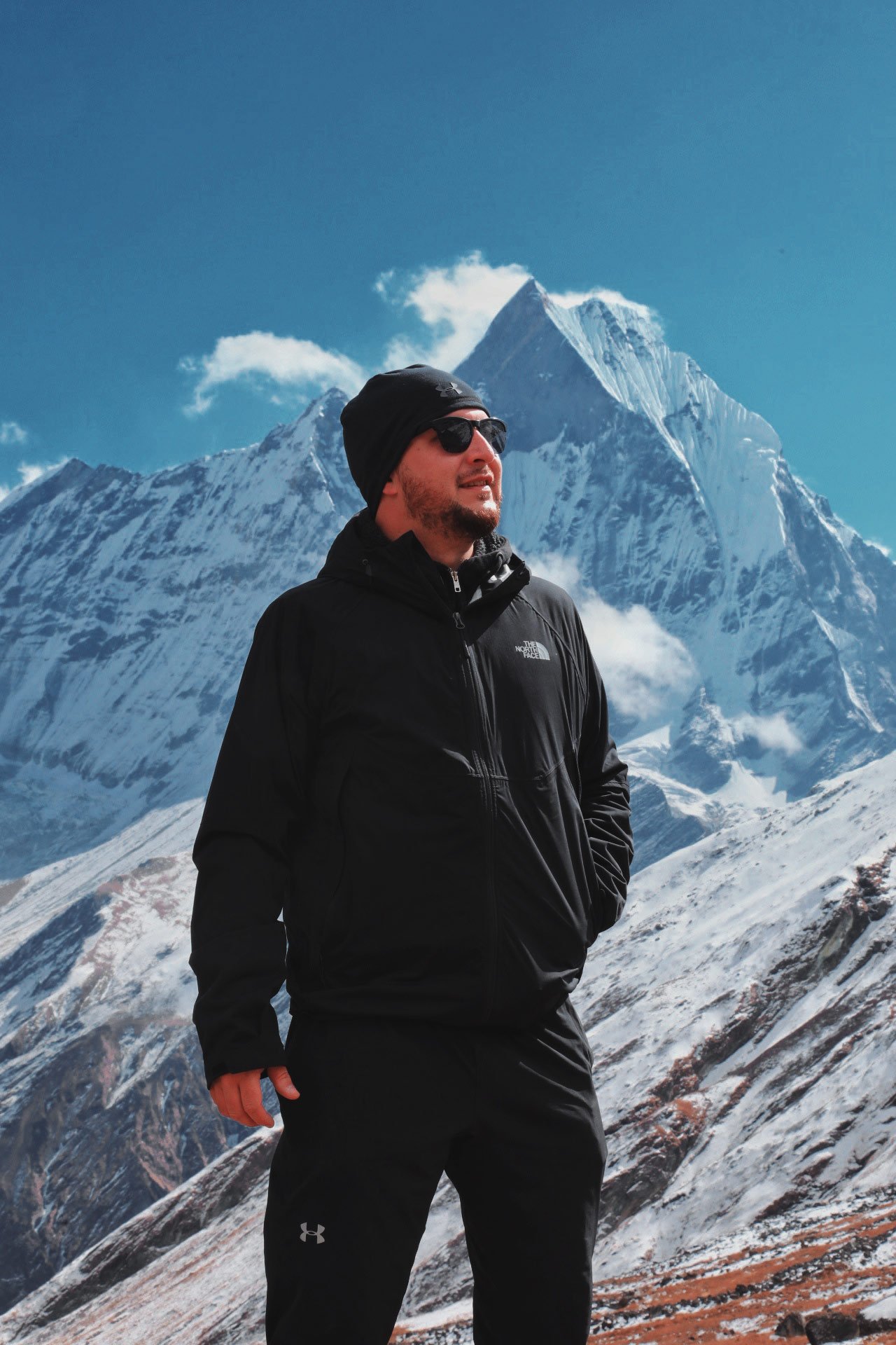 Man dressed in black outdoor gear standing in a snowy mountain landscape with peaks behind him