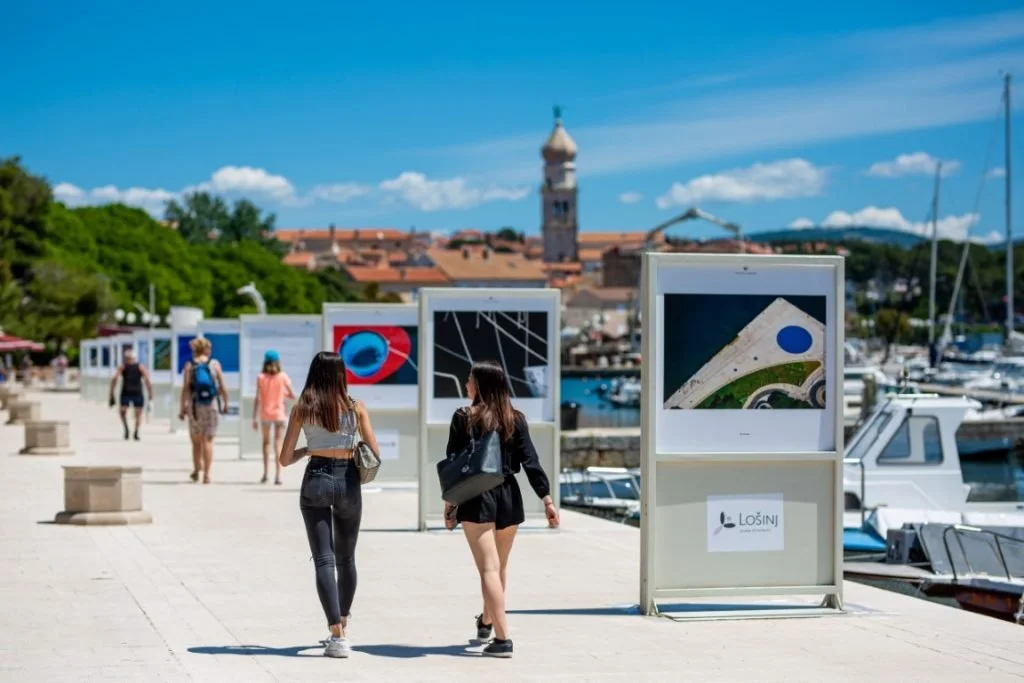 People walking along a waterfront promenade with display boards, boats docked to the side, and a town with a clock tower in the background on a sunny day.