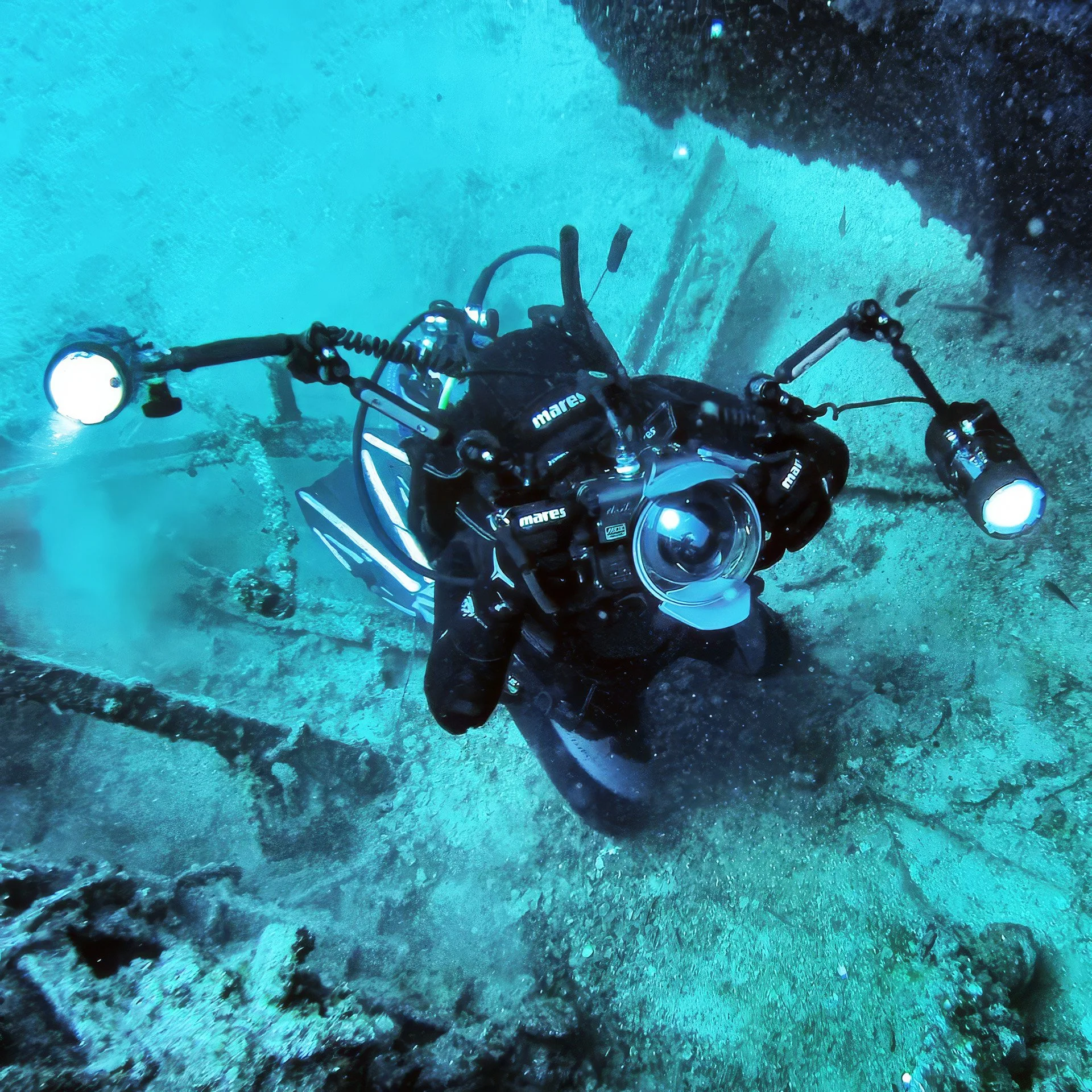 A scuba diver underwater exploring a shipwreck, equipped with a camera and flashlight.