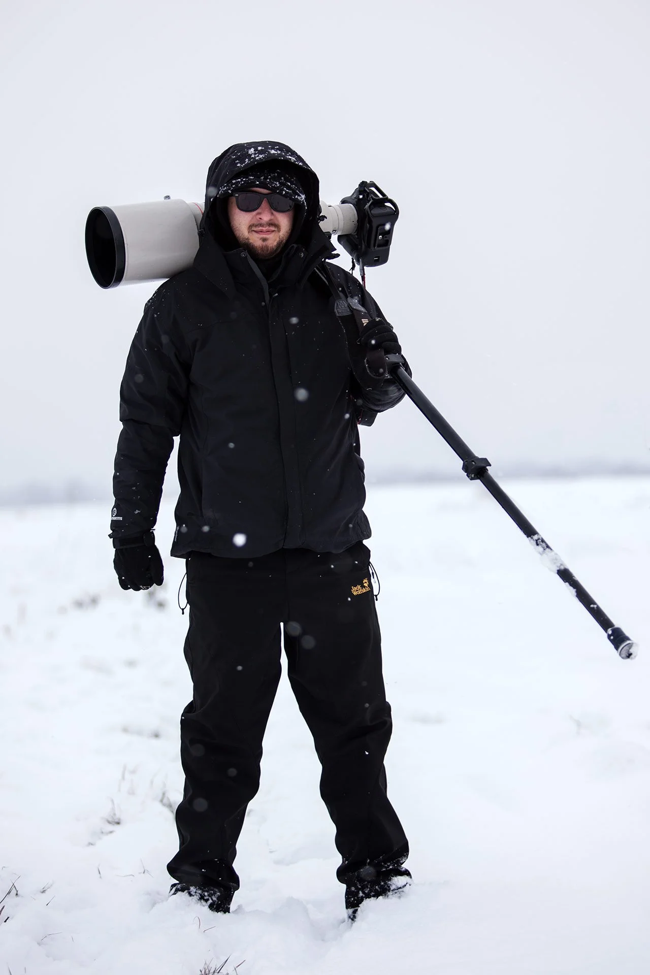 Photographer in black winter gear holding a camera with a large lens on a tripod in a snowy landscape