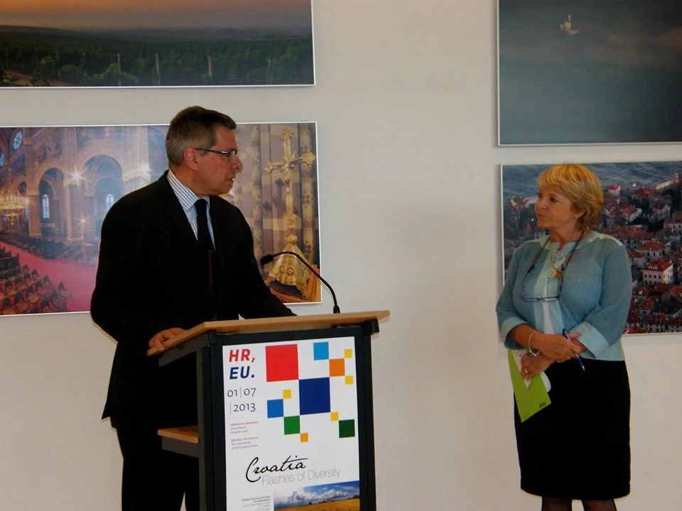 A man in a suit and glasses speaking at a podium with a woman holding a pen and notepad listening. Behind them are framed landscape photos on a white wall.