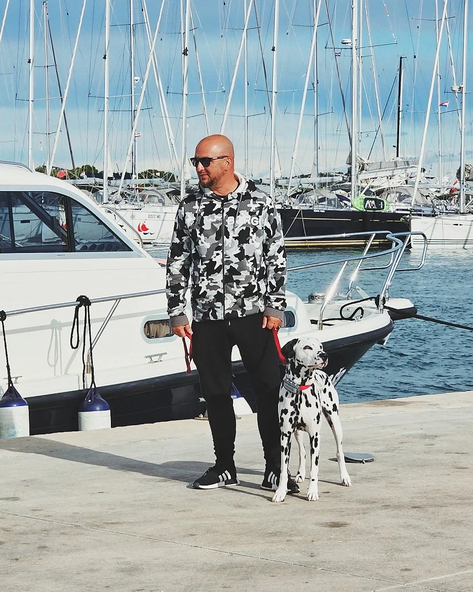A man wearing sunglasses and a camouflage hoodie standing on a dock with a Dalmatian dog, with sailboats and yachts in the background.