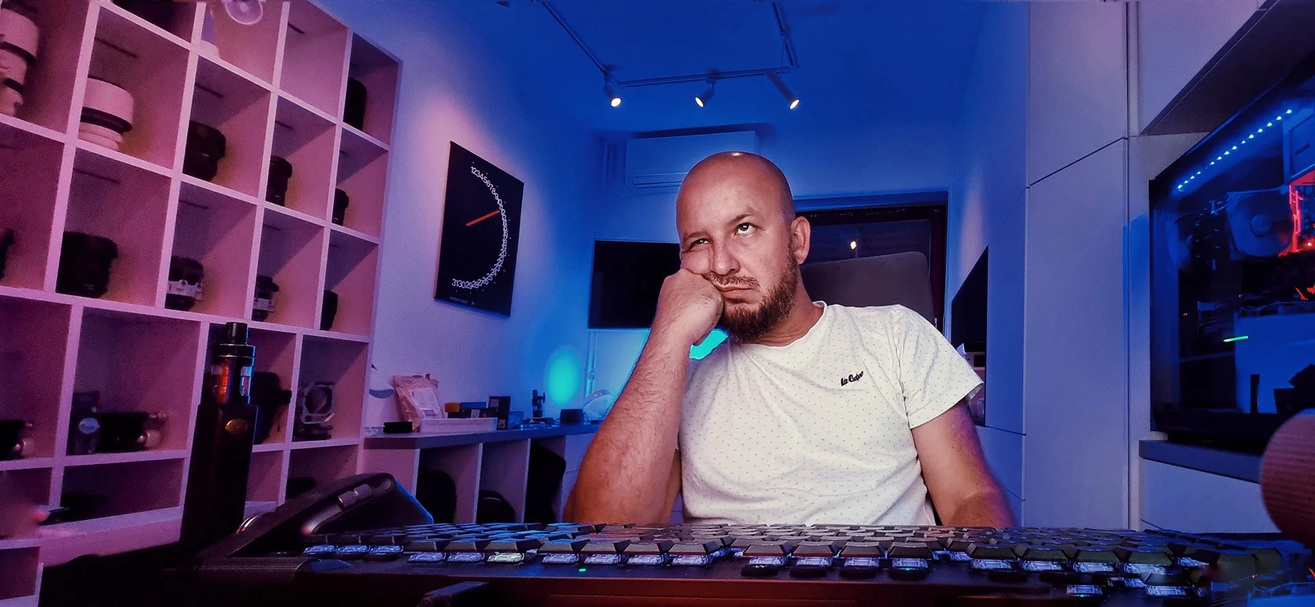 A man sitting at a desk with a computer keyboard, resting his head on his hand, in a room with blue and purple lighting, a wall with cubby shelves filled with jars and objects, and a large monitor in the background.