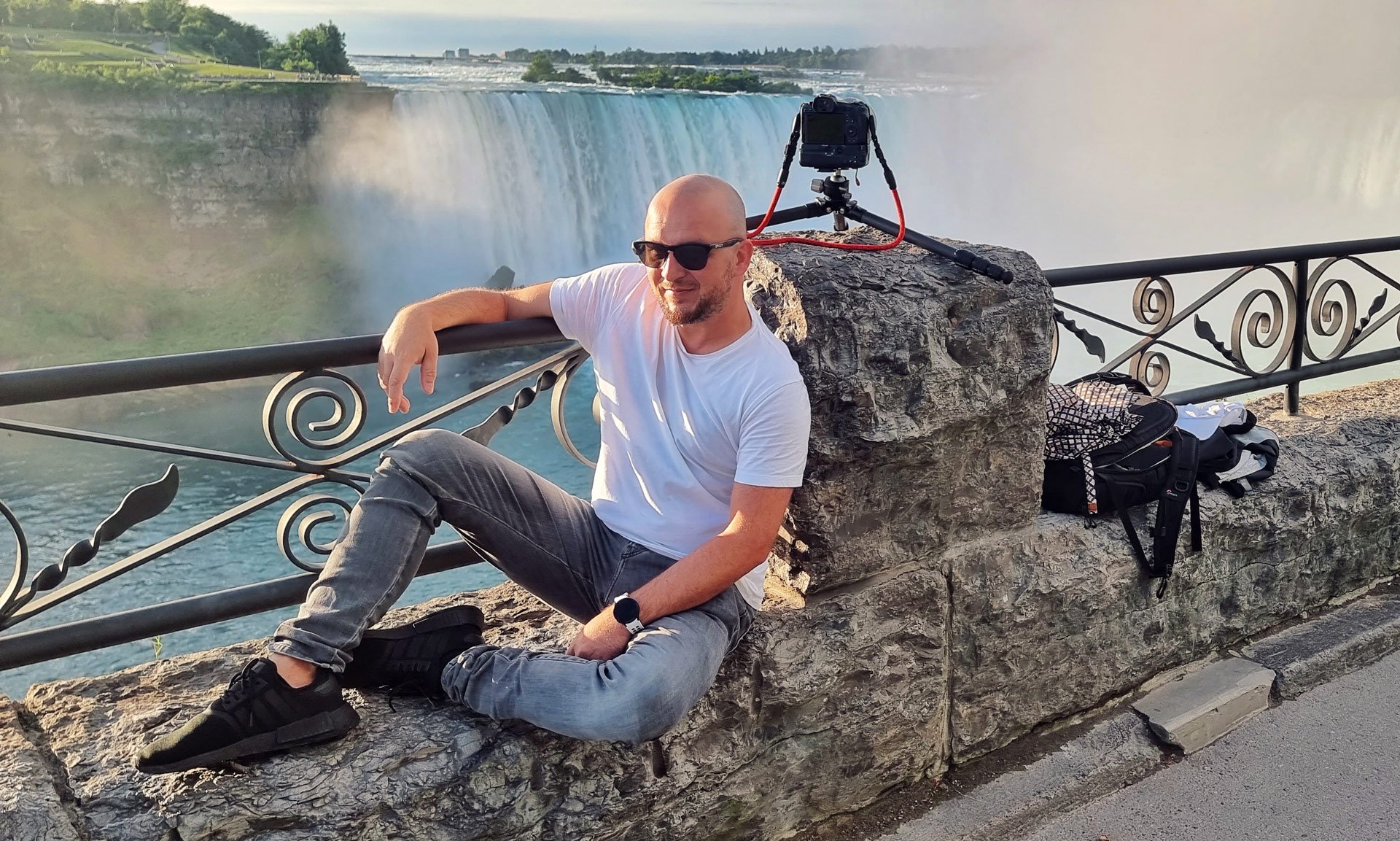 A man wearing a white t-shirt, gray jeans, and black sneakers sitting on a stone ledge near Niagara Falls with a camera on a tripod behind him. He is wearing sunglasses and a black watch, with a backpack and other belongings on the stone beside him. 