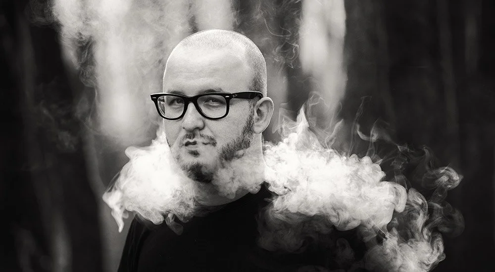A man with glasses and facial hair, outdoors in a forest, blowing smoke that surrounds his face.