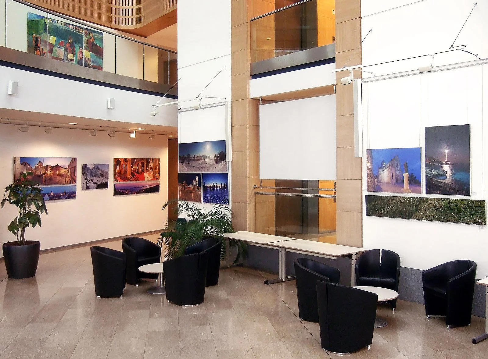 An art gallery or exhibition space with framed landscape and cityscape photographs hanging on white walls. There are six black chairs arranged around small tables, and potted plants are in the corner. The space features a high ceiling and a mezzanine