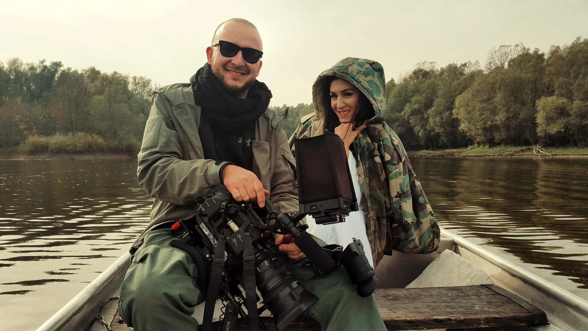 Two people on a boat fishing on a river, one holding a camera and the other wearing a camouflage jacket with a hood.