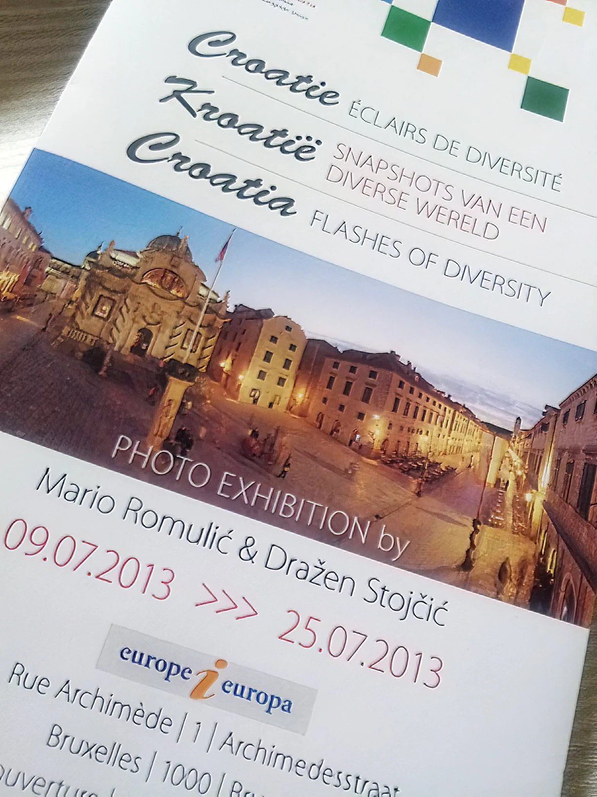 Event poster advertising a photography exhibition titled 'Snapshots of a Diverse World,' featuring images of a historic European city square at dusk, with details about the exhibit dates and location in Brussels, Belgium.