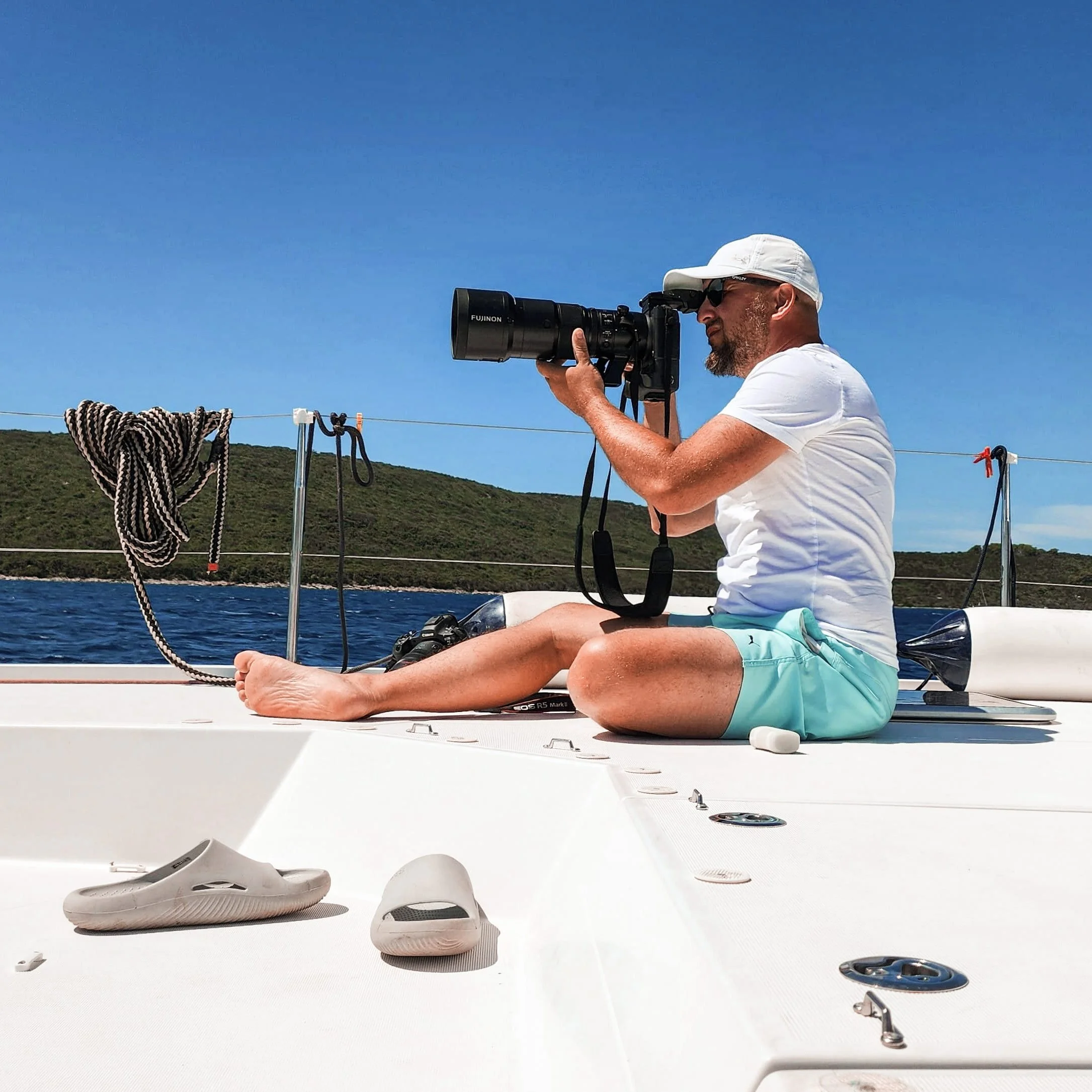 A man sitting cross-legged on a boat deck, taking a photo with a large camera. The background shows a blue sky, water, and a green hilly shoreline.