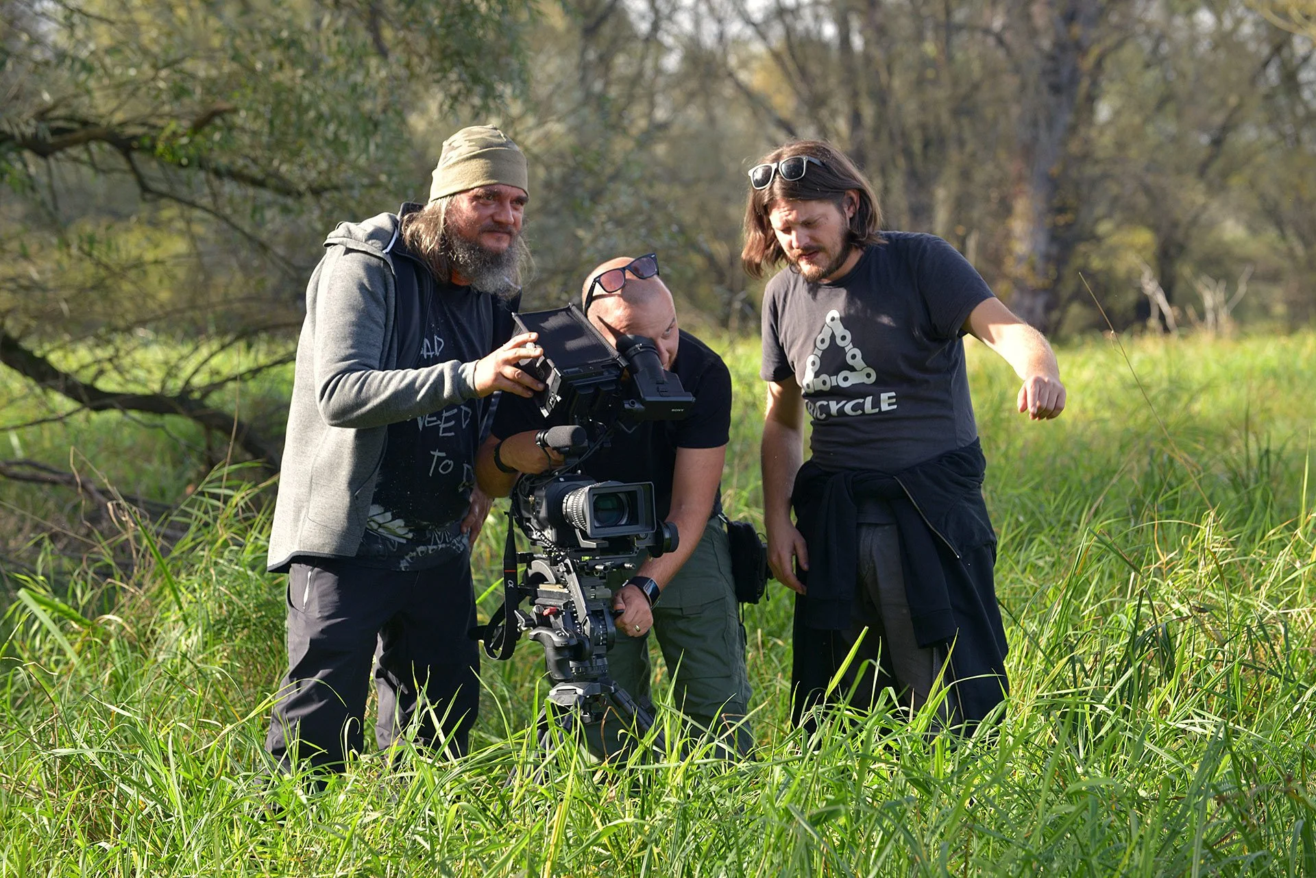 Three men filming outdoors in a grassy field with trees in the background