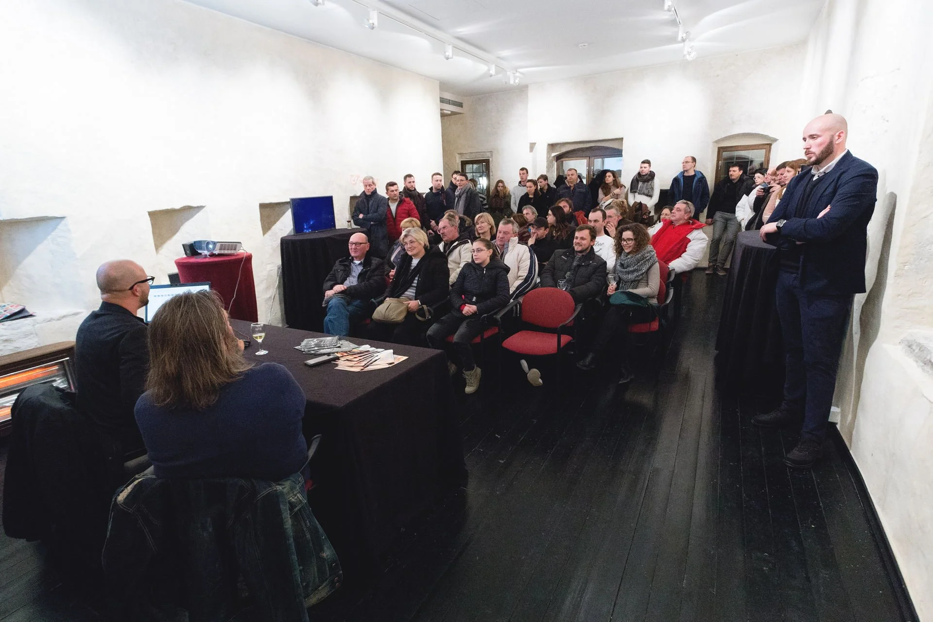 A group of people attending a presentation or event in a room with white walls and dark wooden floors, some seated at tables with computers and drinks, others standing, with a speaker standing on the right side of the room.